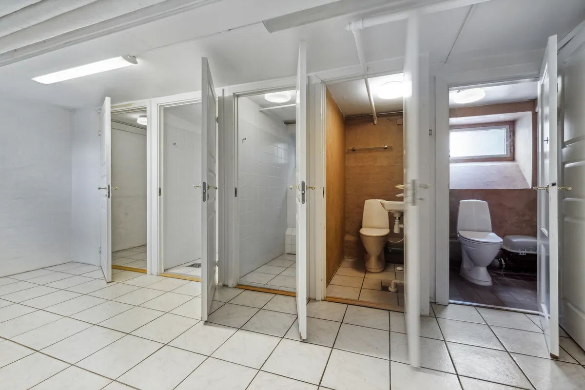 Et stort rum med hvide vægge, hvor der er to toiletter med hvide toilettæpper, og en hvid badeværelseskabinet med hængende lys