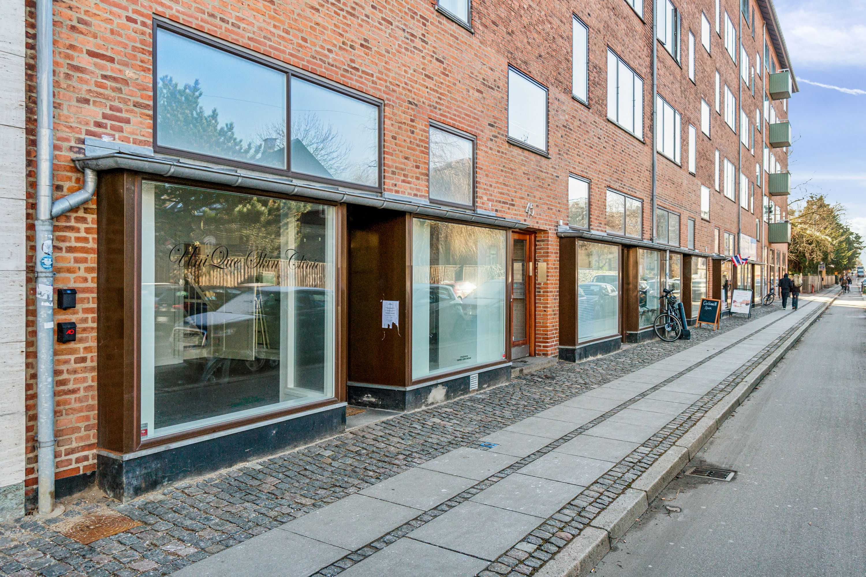 En butik med glasruder og teksten "Vih Que Skin Clinic" på facaden. To mennesker går langs fortovet og en cykel står parkeret. Bygningen har røde mursten og flere lejligheder med balkoner.