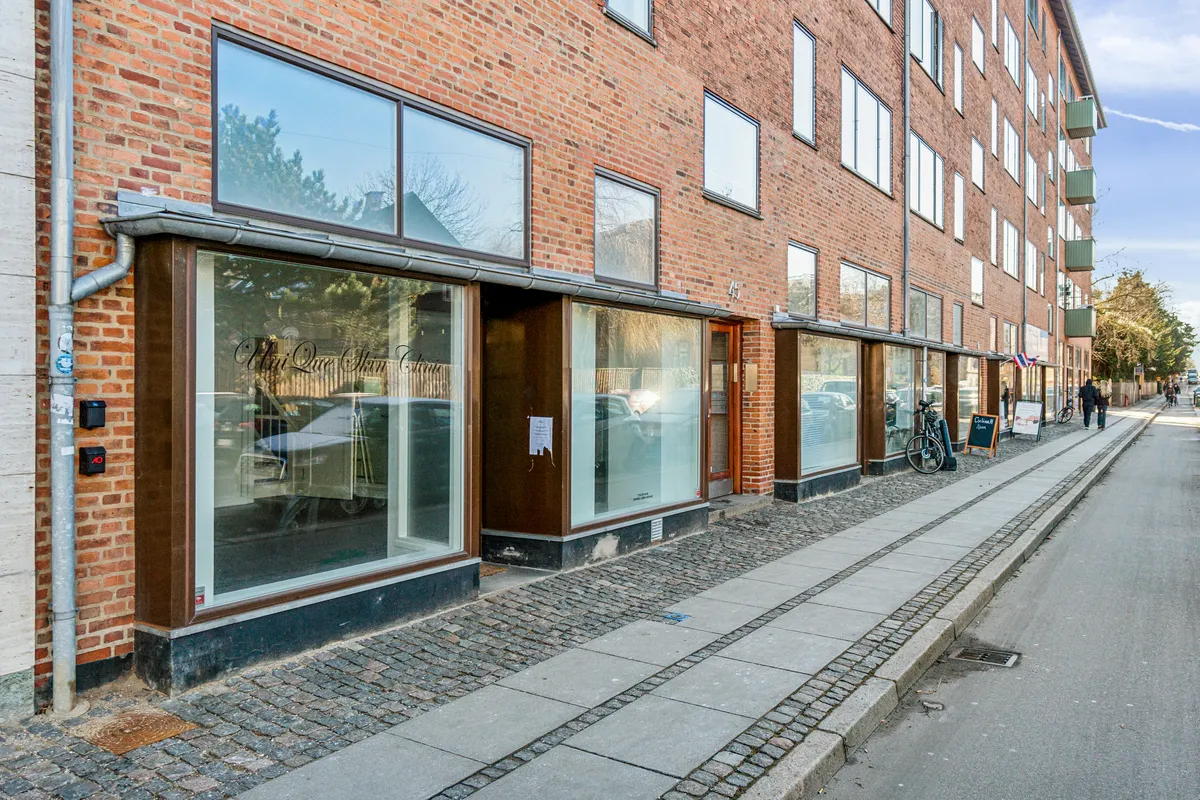 En butik med glasruder og teksten "Vih Que Skin Clinic" på facaden. To mennesker går langs fortovet og en cykel står parkeret. Bygningen har røde mursten og flere lejligheder med balkoner.