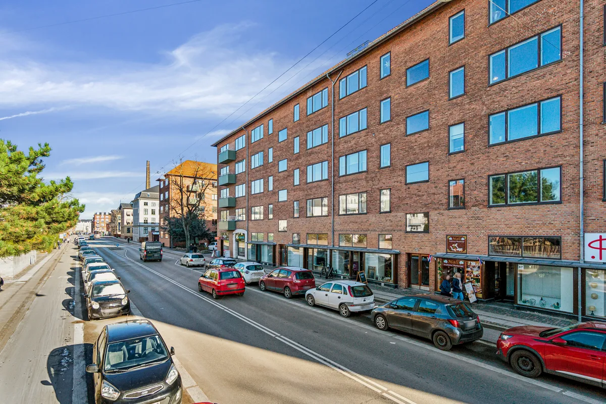 En stor lejlighedsbygning med mange vinduer og køretøjer parkeret på hver side af en bred gade med store træer på begge sider