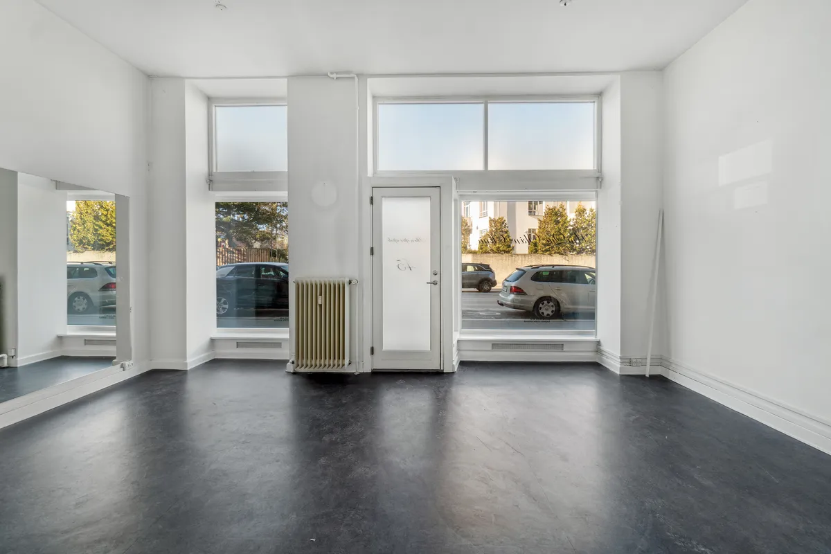 Store butikslokaler med store ruder og lyse indvendige vægge. Væddemål med radiator og gulv med skrald. Udsigt til biler parkeret udenfor og en række træer.