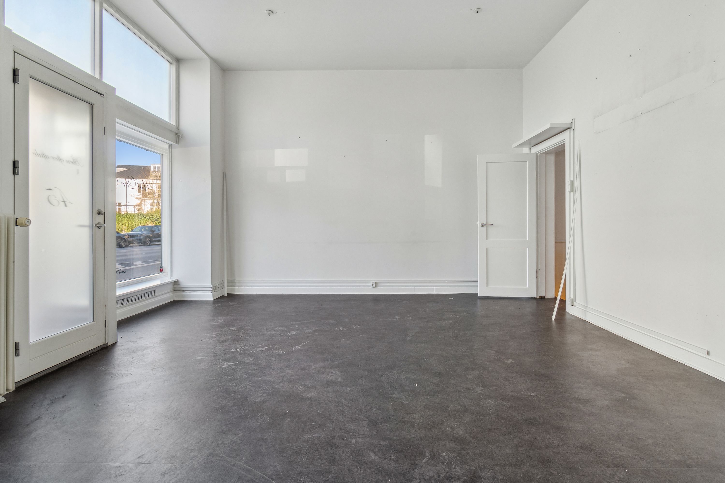 Rummelig butik til leje med højt til loftet og store vinduer. Lysgrå gulv og hvide vægge. To døre med glas og en dør til et mindre rum.