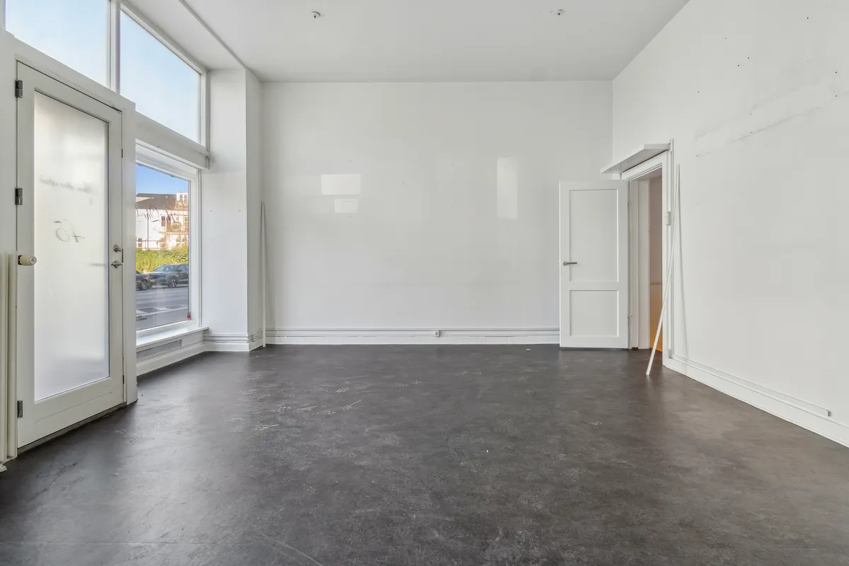 Rummelig butik til leje med højt til loftet og store vinduer. Lysgrå gulv og hvide vægge. To døre med glas og en dør til et mindre rum.