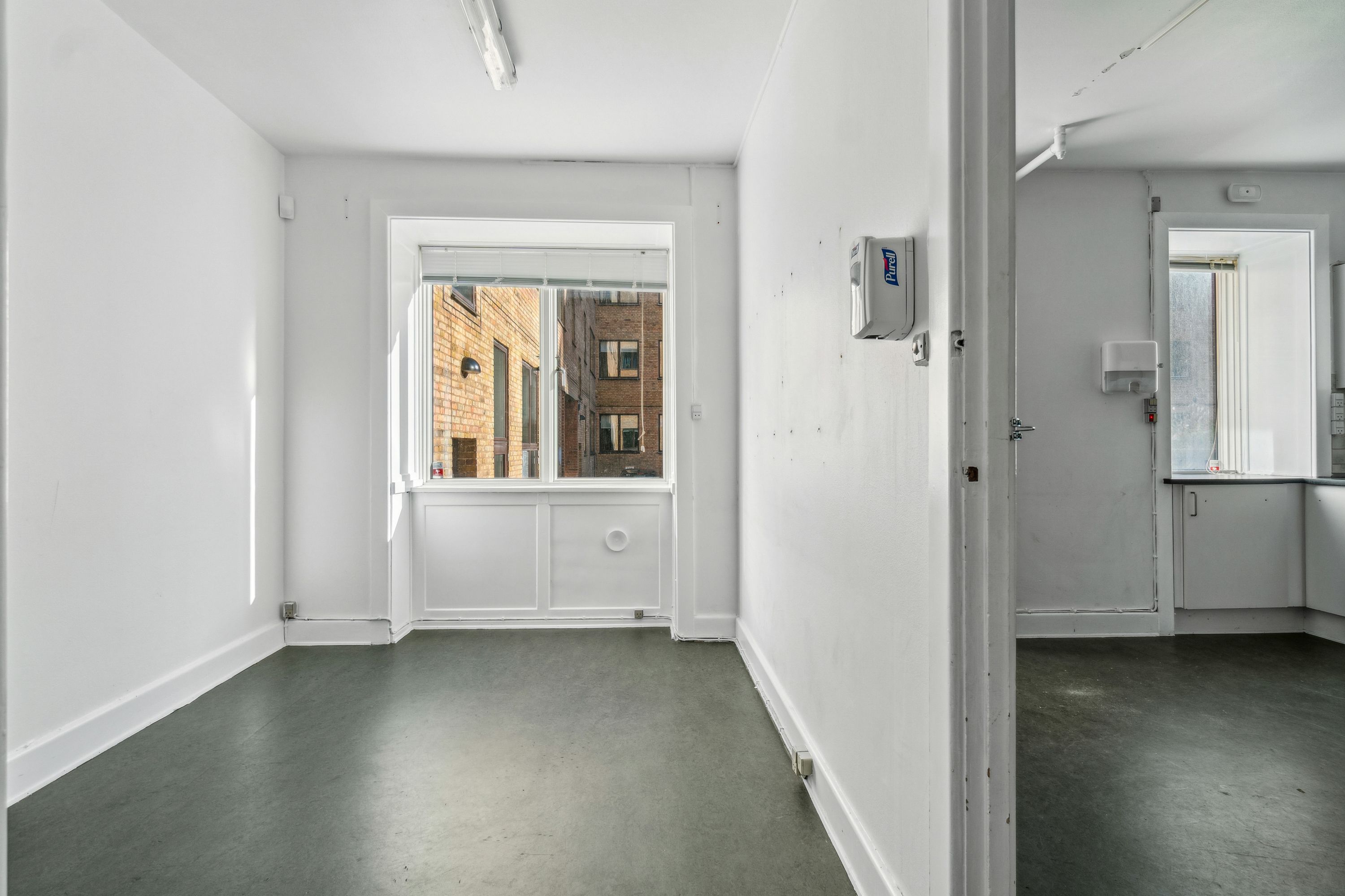 En stor og ren butikslokal med hvide vægge, hvor der er et vindue med udsigt til en bygning med røde mursten og et glasdør. Der er også et hængende lys og en hånddesinficerende enhed på væggen.