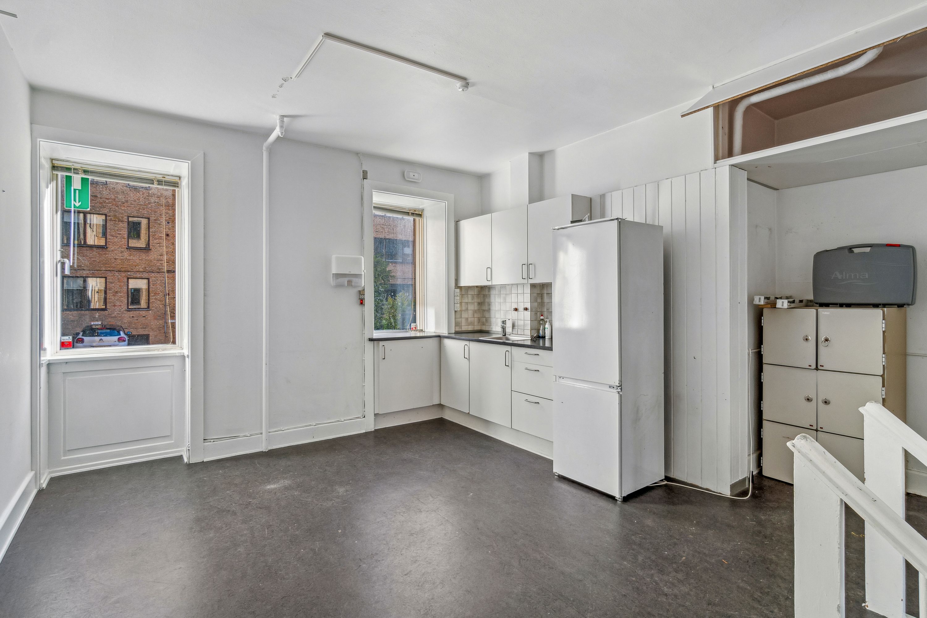 Tom køkken med hvidt køleskab og hvide skabsfronter, tøjtørrer, håndklædetørrer, og vinduesvisning til en bygning med røde mursten og glasruder