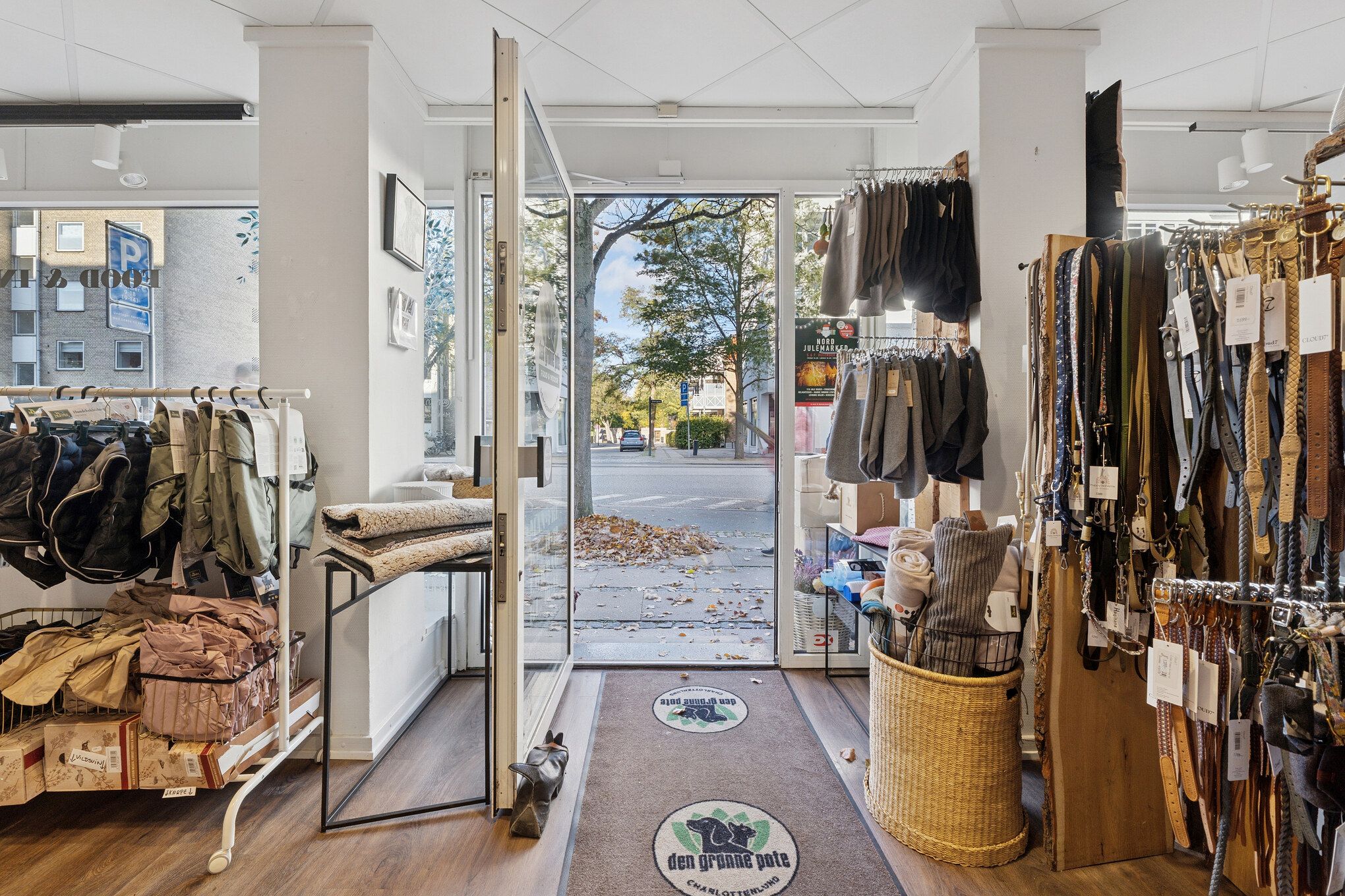 Lyst og indbydende butikslejemål med god eksponering og fleksibel indretning. Bred facade med to store vinduer og indgangsdør. Sorte og brune stoffer, smykker og tøj er udstillet. Fortov og biler ses i baggrunden.