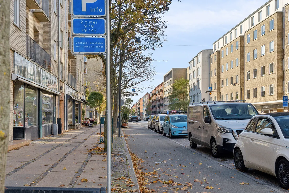 Parkerede biler og et infoskilt for bilparkering i Charlottenlund. I baggrunden ses en bygning med flere lejligheder og en gågade med butikker og terrasser.