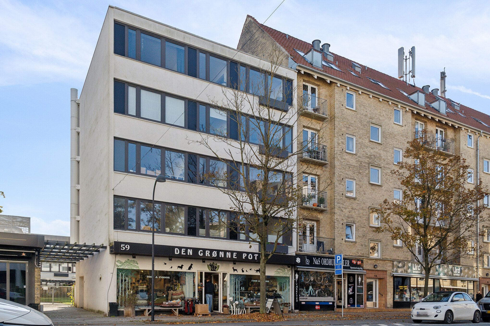 Menighedsbygning med butik Den Grønne Pot, to biler parkeret, træer og kørestolsvenlige fortov, lyse himmel, flere butikker og et højhus med røde tag og mange vinduer