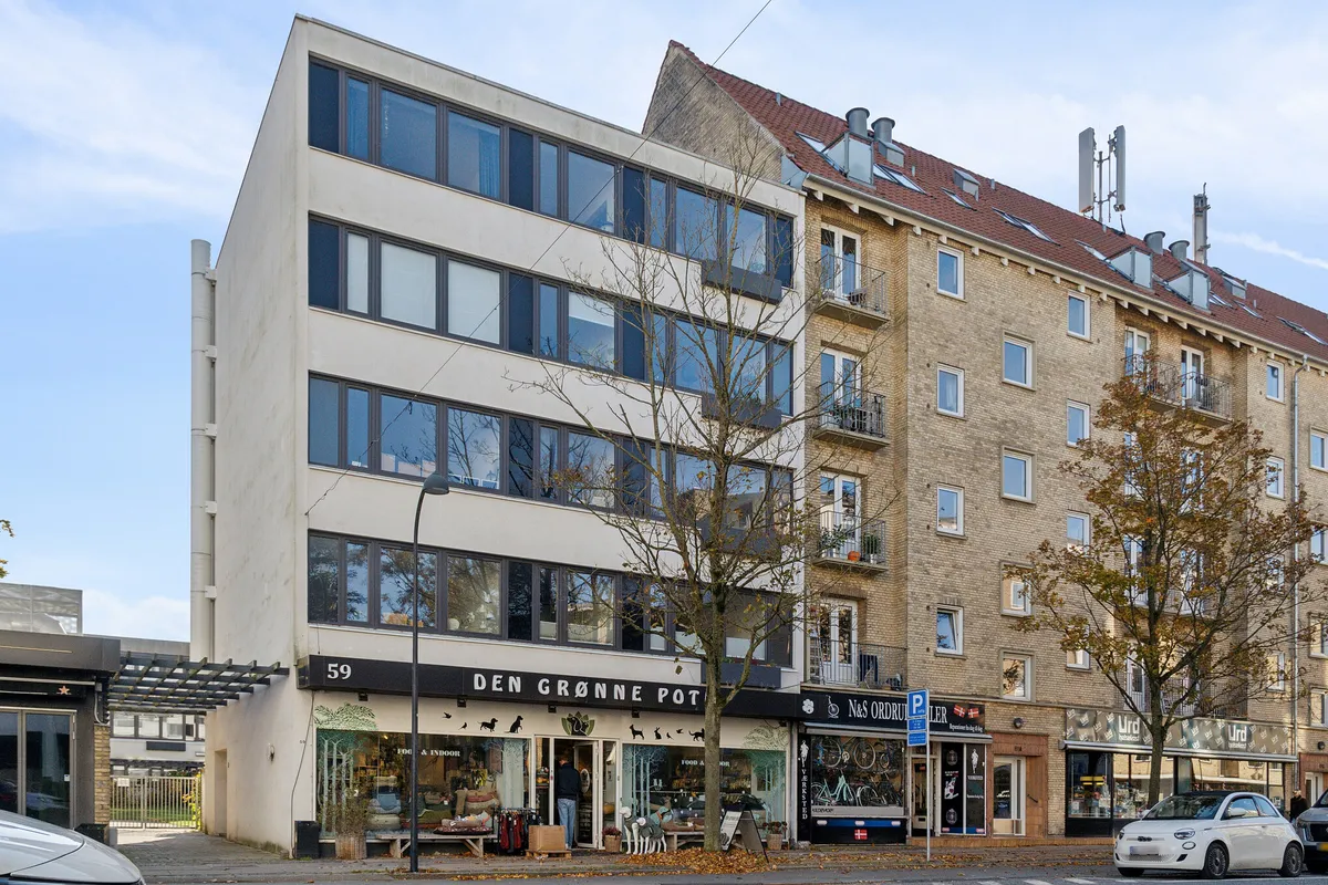 Menighedsbygning med butik Den Grønne Pot, to biler parkeret, træer og kørestolsvenlige fortov, lyse himmel, flere butikker og et højhus med røde tag og mange vinduer