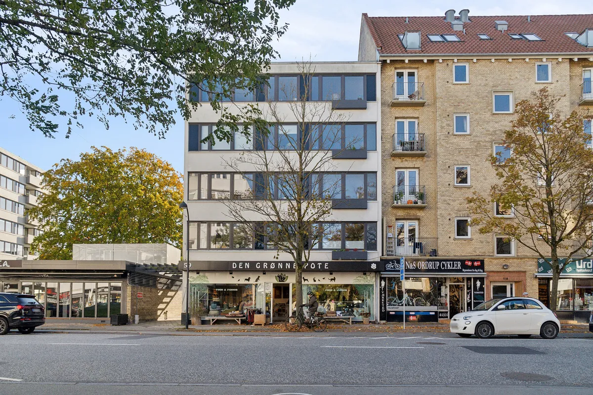 Menighedsgårdens facade med butikker, hvor Den Grønne Pote ligger, og en cykelforretning til højre. En mand kører på en cykel foran den grønne facade