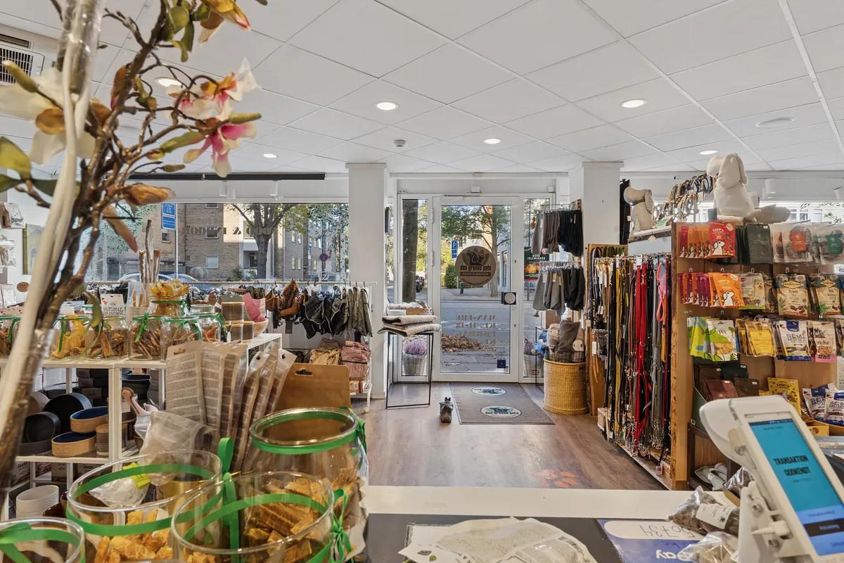 Butik med varer til dyr, herunder foder, senge og legetøj. Inde er der et kassebord med kassen og en plante med blomster. Butikken har trægulv og lyse lofter med rundt rundt.