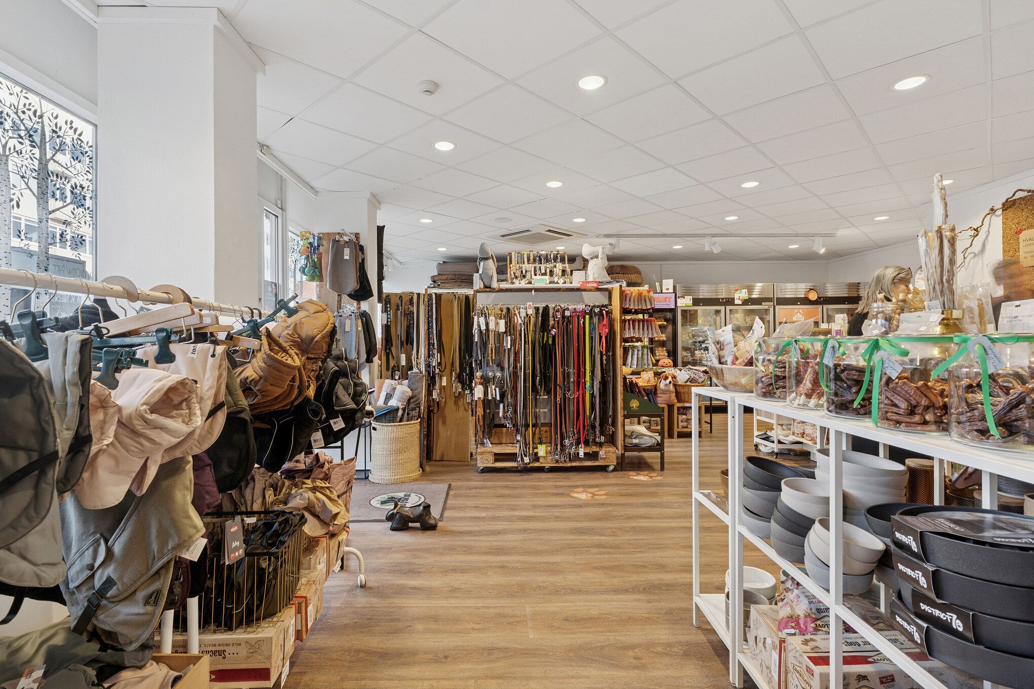 Lyst og indbydende butikslejemål med mange produkter, inkluderet tøj, tasker, legetøj, sko og glasbeholdere med snacks. Dekorative gulvmotiver og lyse loftslamper skaber en hyggelig atmosfære