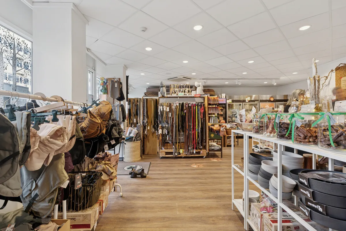 Lyst og indbydende butikslejemål med mange produkter, inkluderet tøj, tasker, legetøj, sko og glasbeholdere med snacks. Dekorative gulvmotiver og lyse loftslamper skaber en hyggelig atmosfære