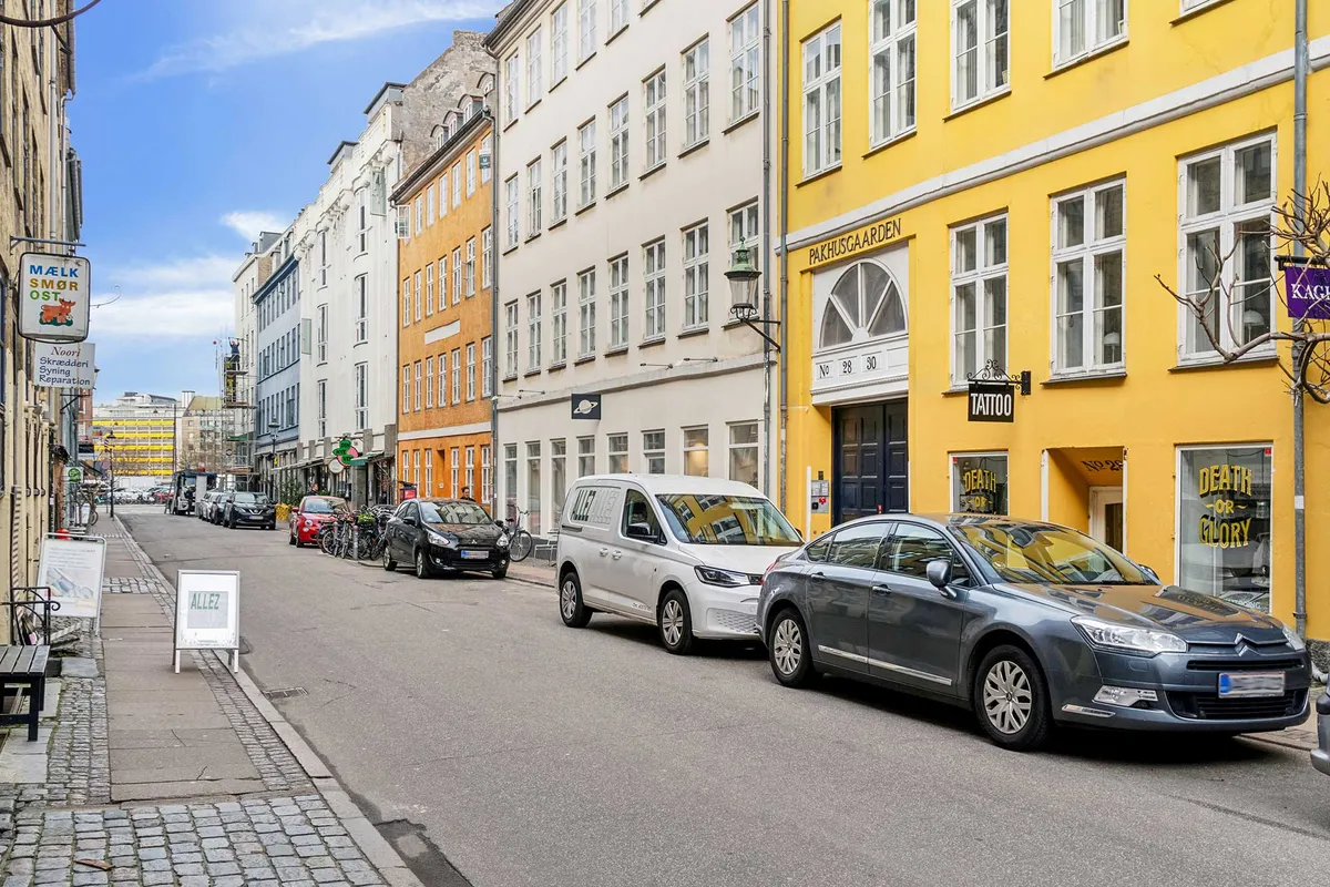 To biler parkeret på en gade i København, foran en butik med skiltet "TATTOO" og "DEATH OR GLORY". På fortauet er der en skilt med teksten "ALLEZ" og en række bygninger i baggrunden.