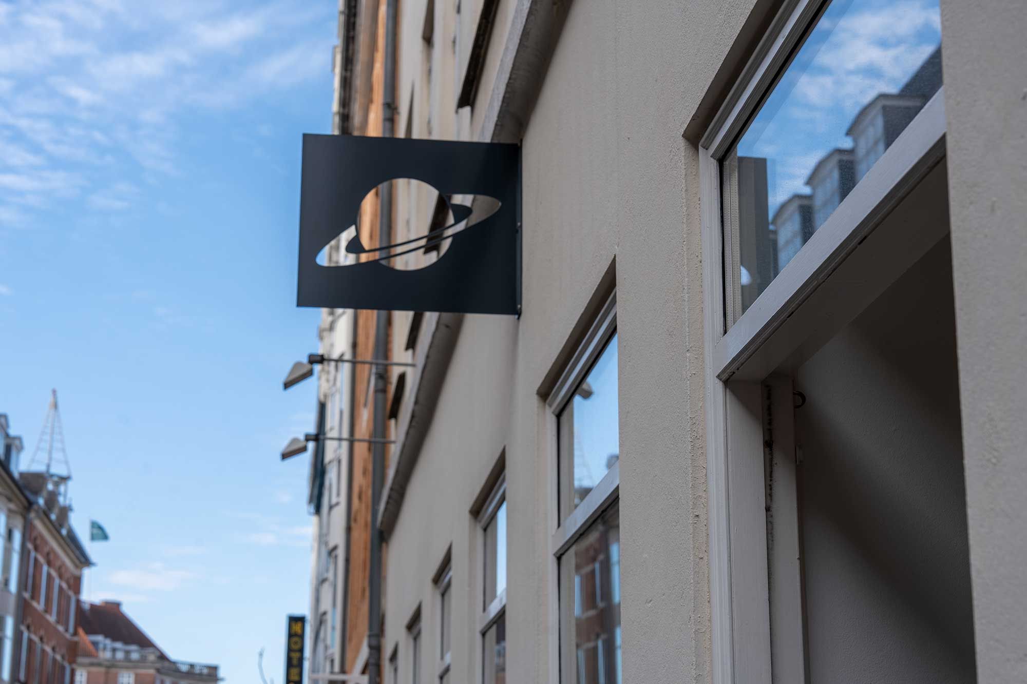 Markant butikslejemål med Saturn-logo i Latinerkvarteret. Lejemålet ligger på en hjørnebygning med store vinduer og refleksioner i glas. Skyen er lyseblå med lyseregn.