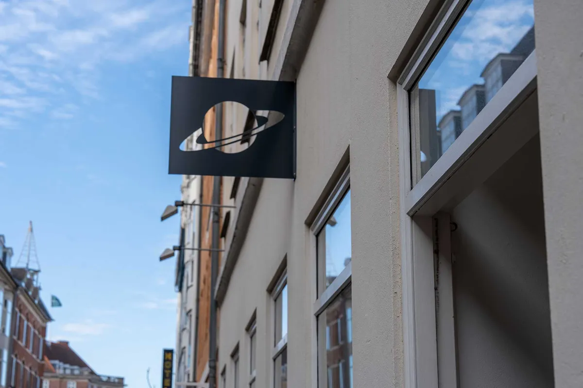 Markant butikslejemål med Saturn-logo i Latinerkvarteret. Lejemålet ligger på en hjørnebygning med store vinduer og refleksioner i glas. Skyen er lyseblå med lyseregn.