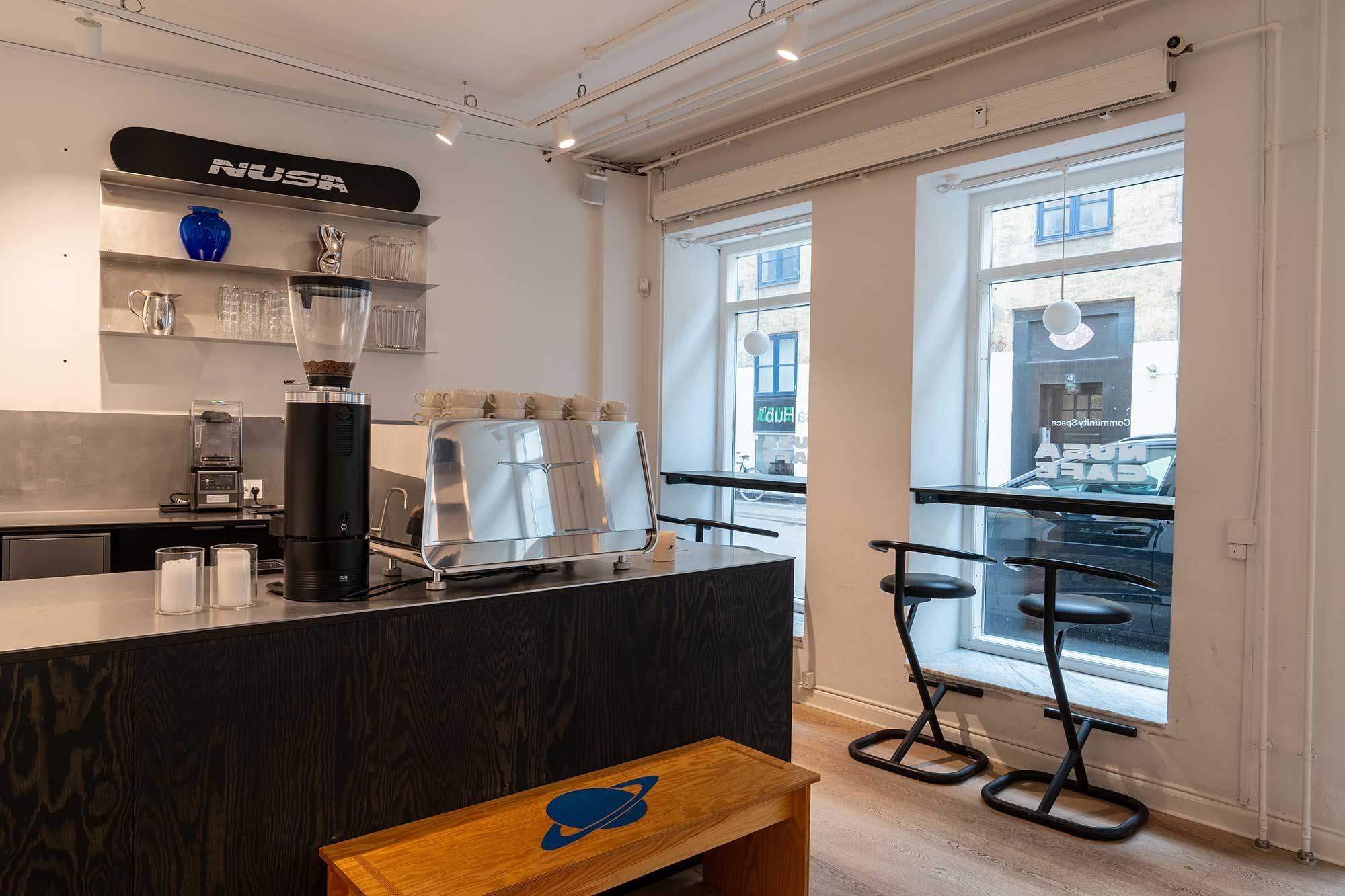 En køkkendel i et lejemål med lyse loftslampe og eksponeret kaffemaskine og sølvgrej. Der er to høje stole og et bord med et køretøj udenfor vinduet.