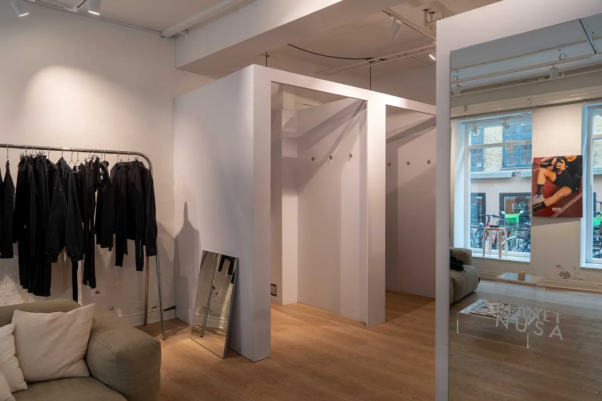 Et moderne indretningsstudie med lyse vægge og højloft samt store spejl. Tøj og møbler er placeret i rummet, og der er en udsigt til en bygning med cykler udenfor.