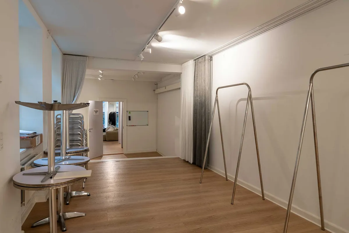 Lejlighedslejemål i Latinerkvarteret med salgsområde, garderobestandere, flere bordopstativeringer og åben dør til bagværelse med skrivebord og sofa