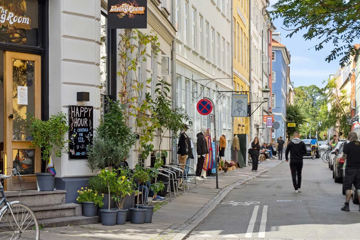 Mennesker går på gågaden i Latinerkvarteret og nyder københavnsvejret. Butikken med cocktail-menuen står åben, og der er planter og stole på fortauet