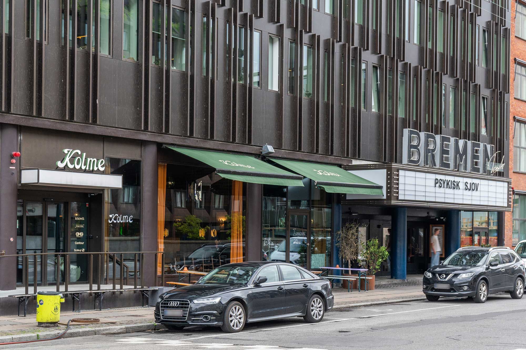 To sorte biler er parkeret udenfor et bygningskompleks med et kinesisk navn og en cafe med grønne kulisserne