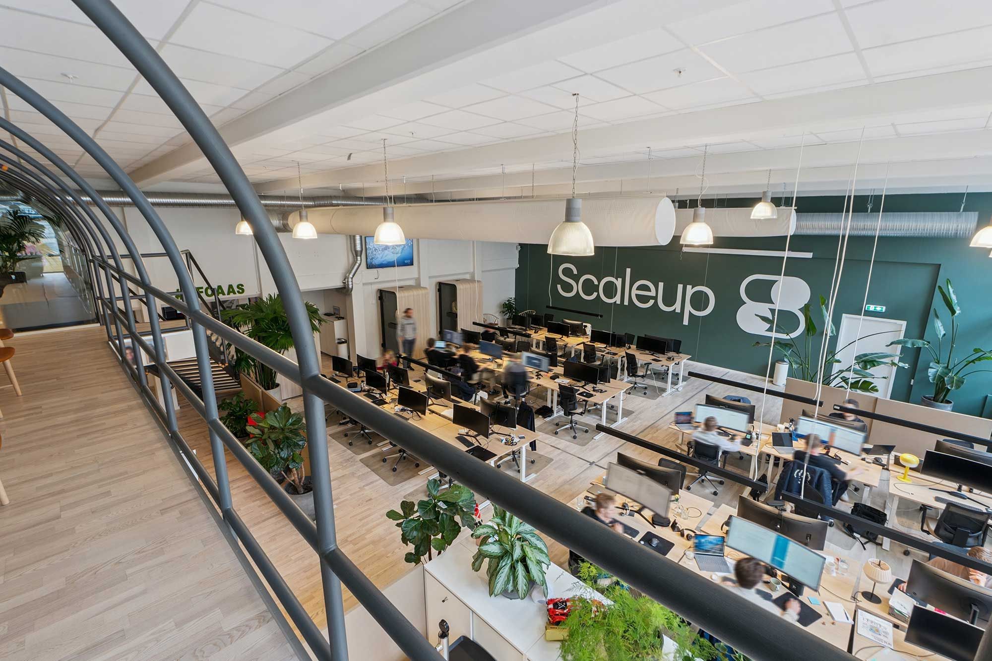 Kreativt kontor med åbne områder, plantegrønne og højt til loftet. Flere medarbejdere arbejder på computere med monitorer, og kontoret er opdelt i flere områder med møbler og teknologi.