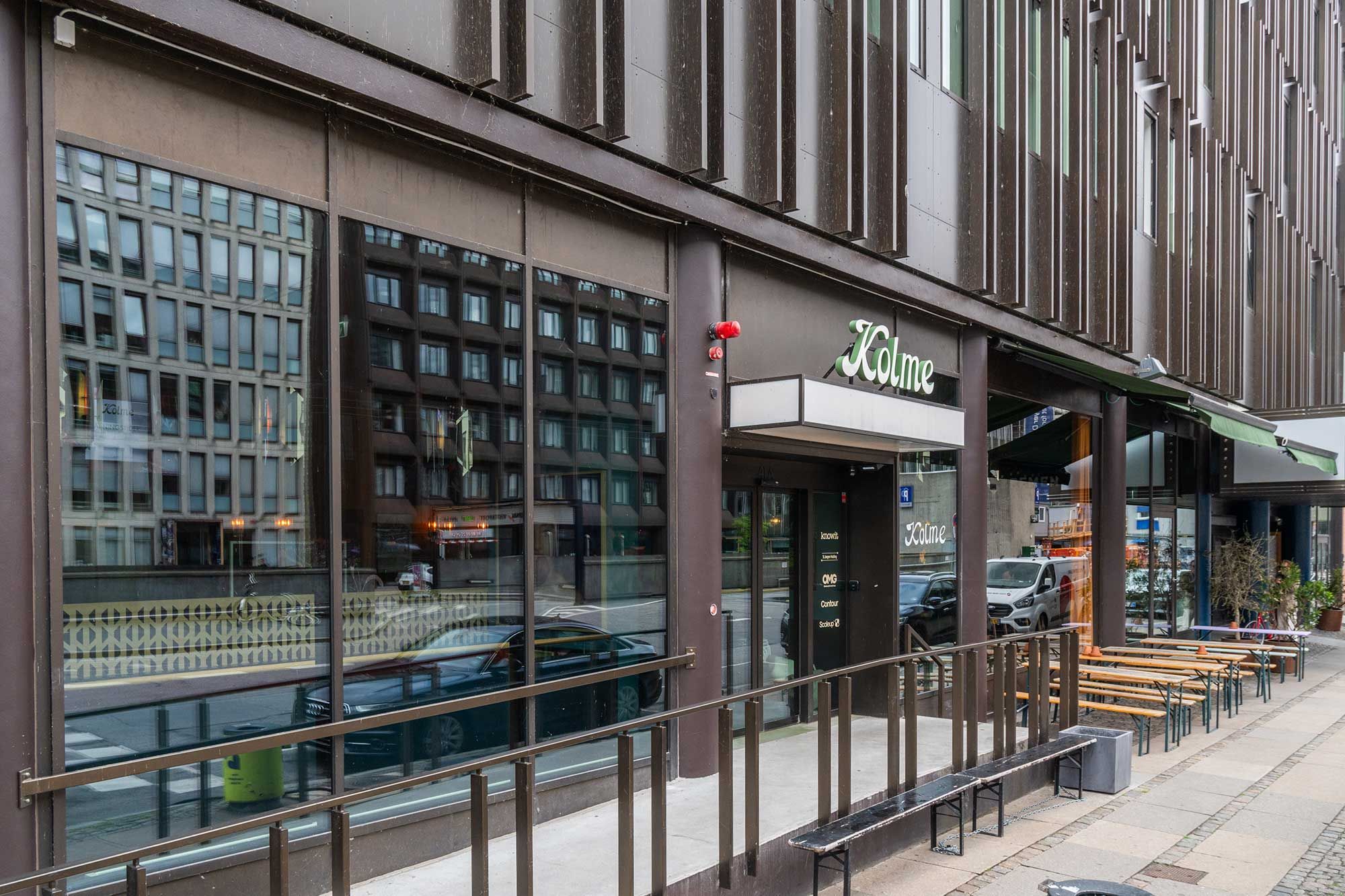 Moderne kontorbygning med store vinduer og glasdøre, terrasse med bænke og stole, vej og biler i baggrunden, grønt plante i hjørnet, skrift "Kolme" på facaden