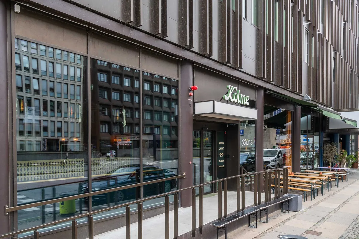 Moderne kontorbygning med store vinduer og glasdøre, terrasse med bænke og stole, vej og biler i baggrunden, grønt plante i hjørnet, skrift "Kolme" på facaden