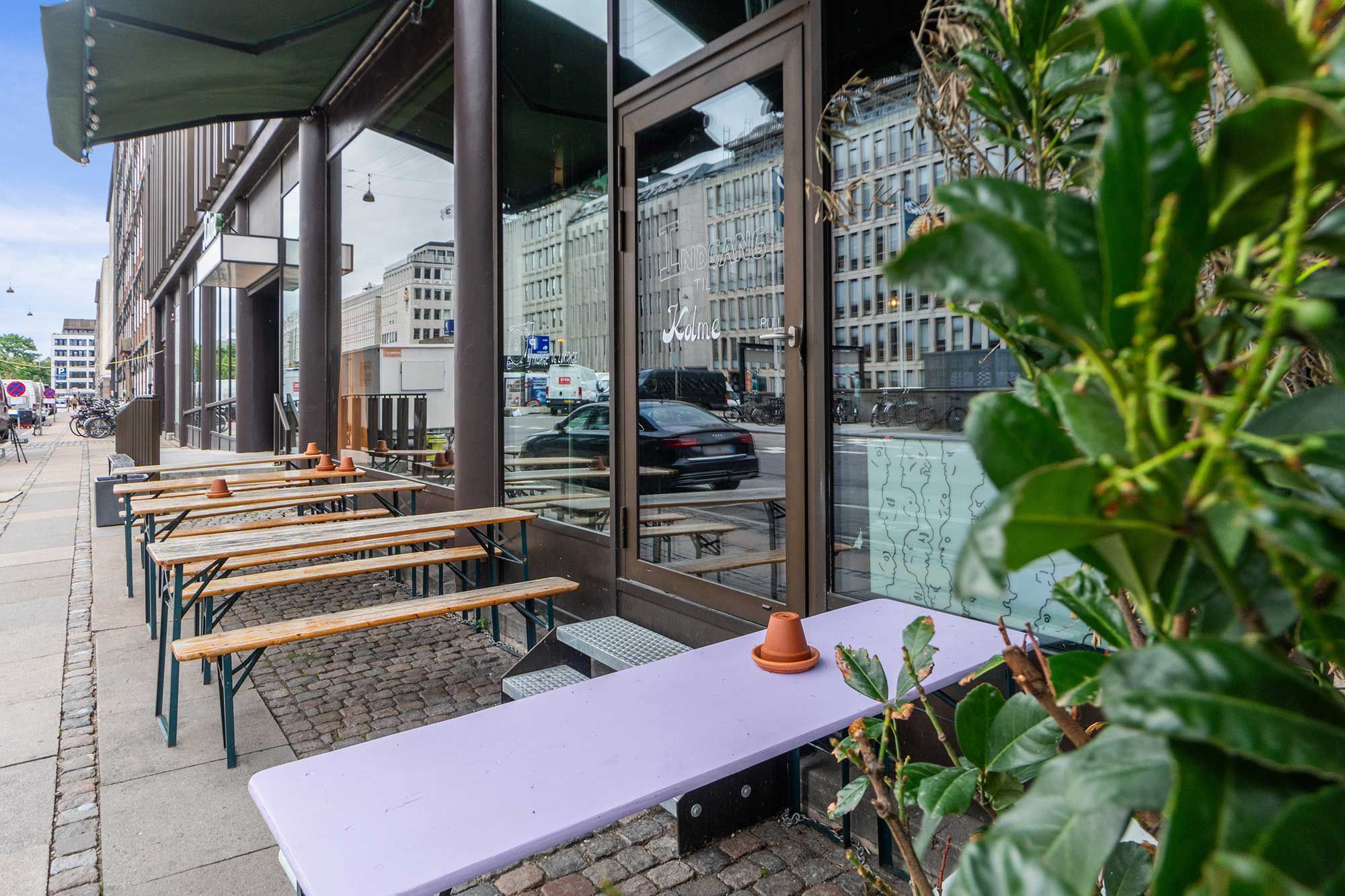 Lille restaurant med udsigt til gaden, hvor der er parkerede biler og cykler. En plante med grønne blade og brun gryde står på en lyslilla lang stol.