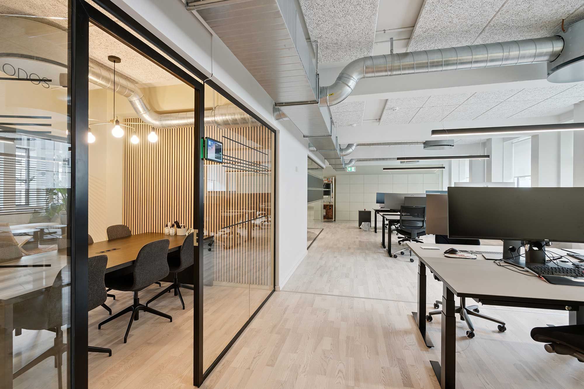 Moderne mødeværelse med computere og skriveborde, højt gulv og hvide vægge. Værelse med lyse lysekroner og træindretning langs væggen. Forskudte stolpe og højt til loftet.
