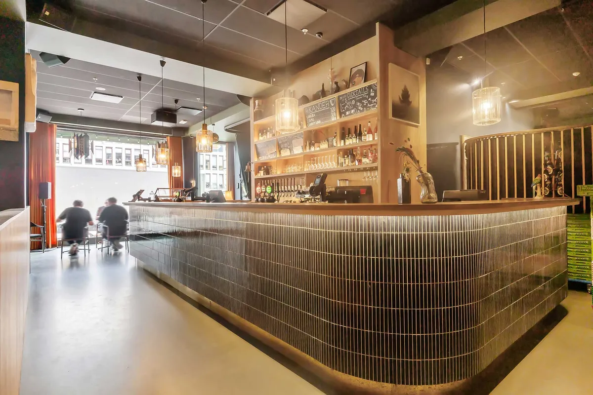 Inde i en café med rustfri stål og højt gulv, to mænd sidder ved borde og nyder drikkevarer. Lys pære og dekorative vinflasker på hylden og lysekroner hængende fra loftet