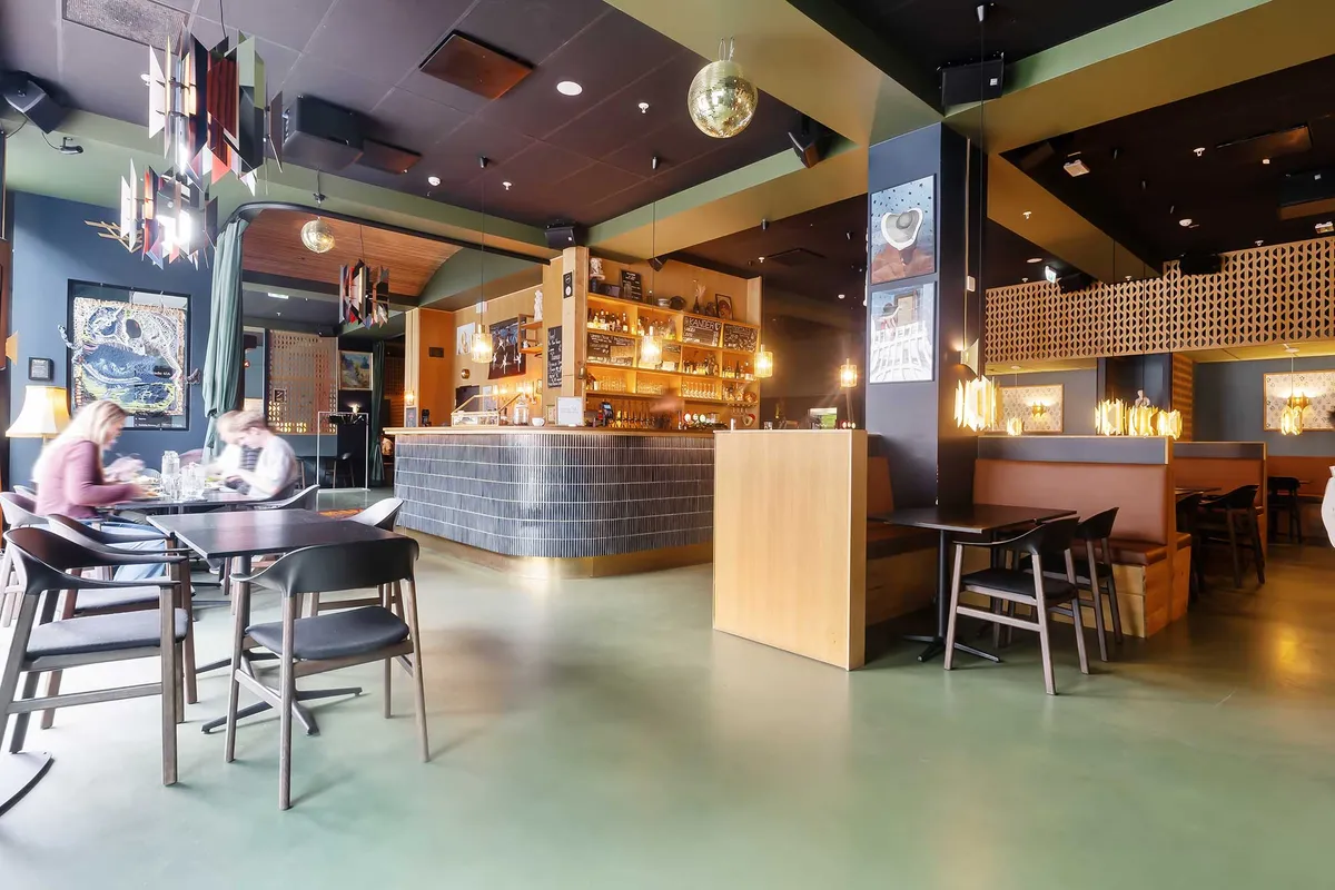 Interiør med åbent plan, lyse farver og flotte lysekroner. Rundt om er der små bord og stole samt en bar. Der er to kvinder ved bordet, der spiser sammen.