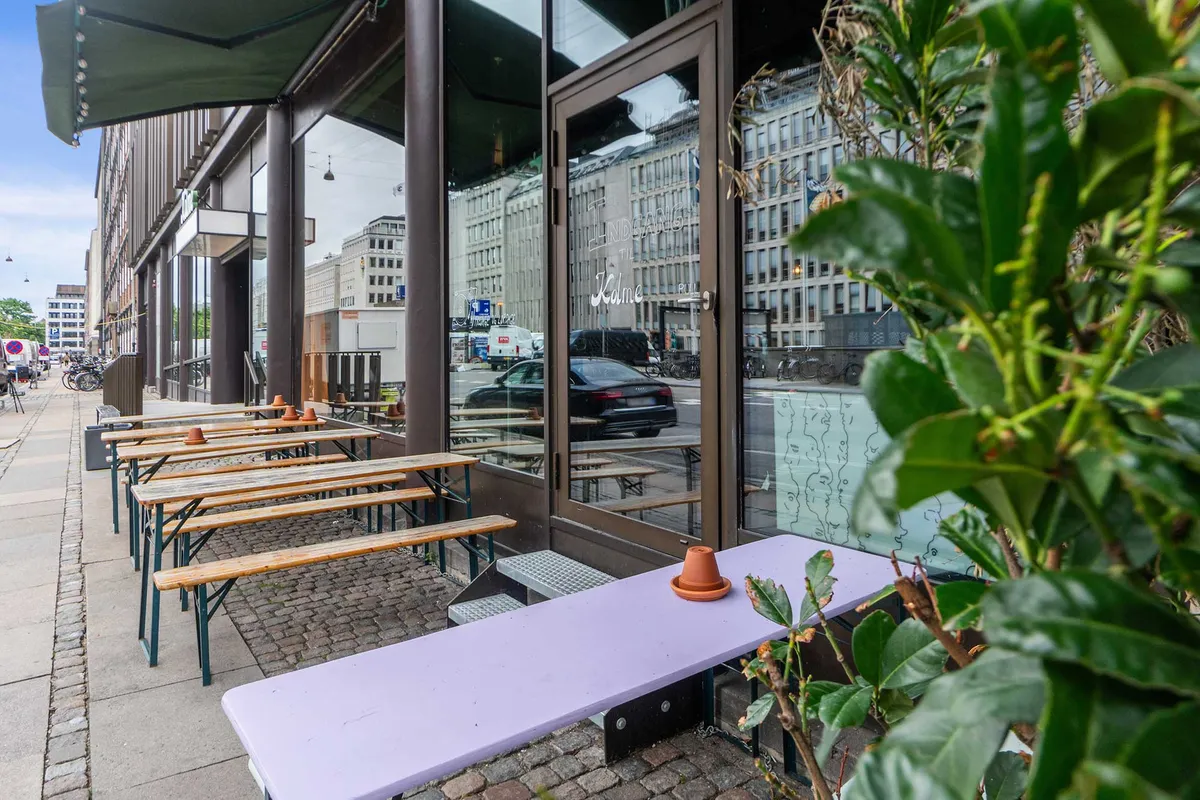 En lille terrasse med træborde og bænke i lyse farver foran et cafévindue med lysereflekser fra biler og bygninger på baggrunden.