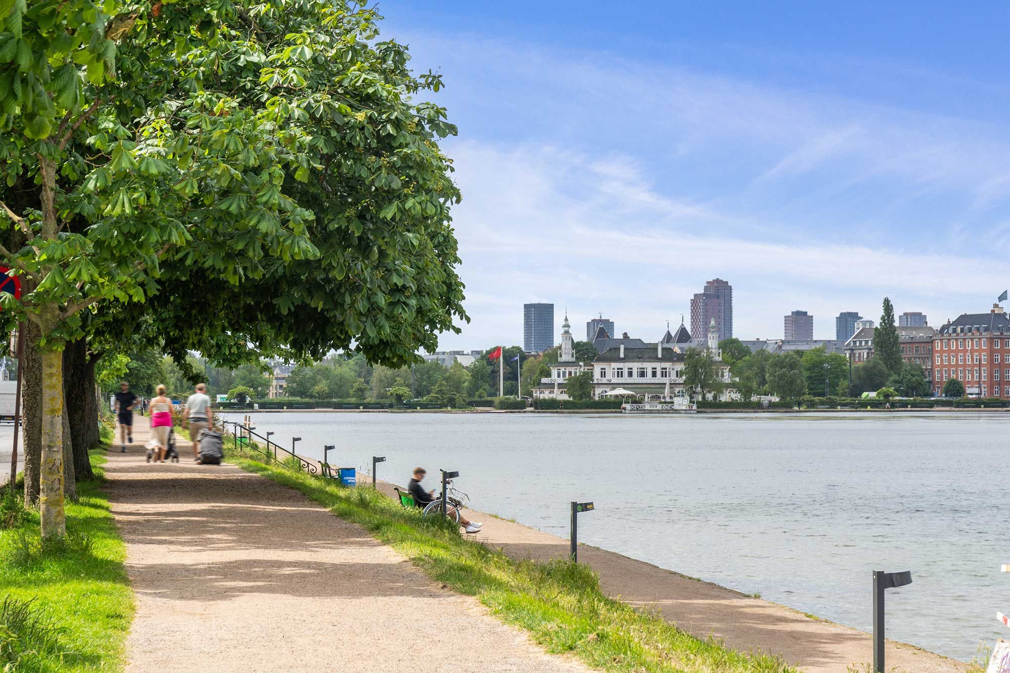 Mennesker går på en promenade langs vandet med bygninger i baggrunden, der har tårne og flag