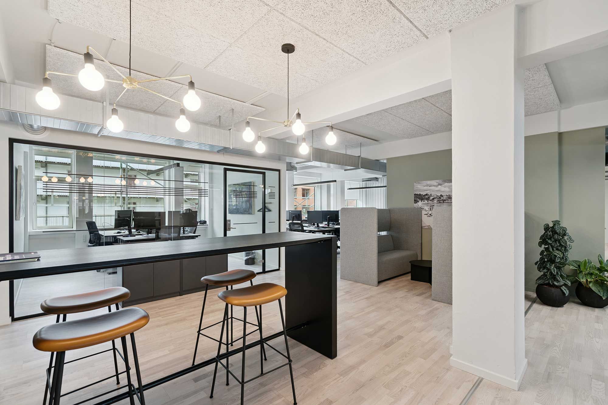 Lys indrettet kontor med trægulv og lysekroner med lyseperler hængende fra loftet. Kontorbord med fire lædersæde stole samt kontormøbler og plantager i en væg.