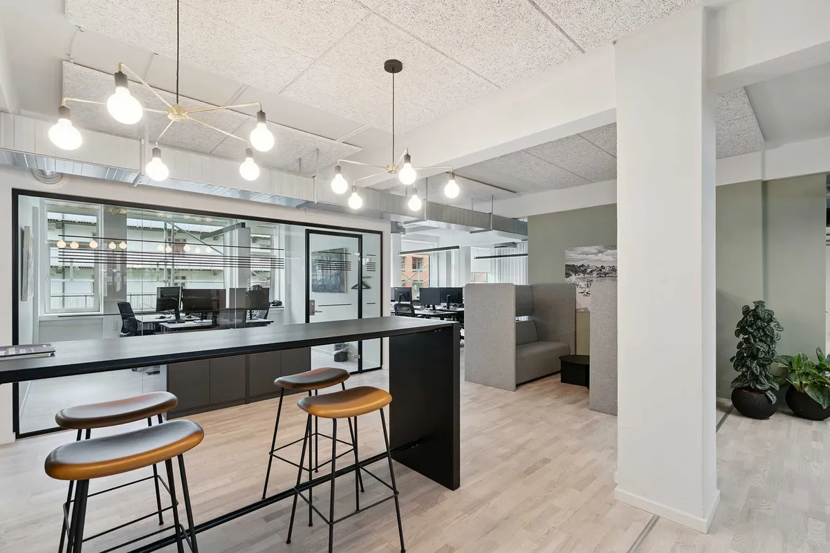 Lys indrettet kontor med trægulv og lysekroner med lyseperler hængende fra loftet. Kontorbord med fire lædersæde stole samt kontormøbler og plantager i en væg.