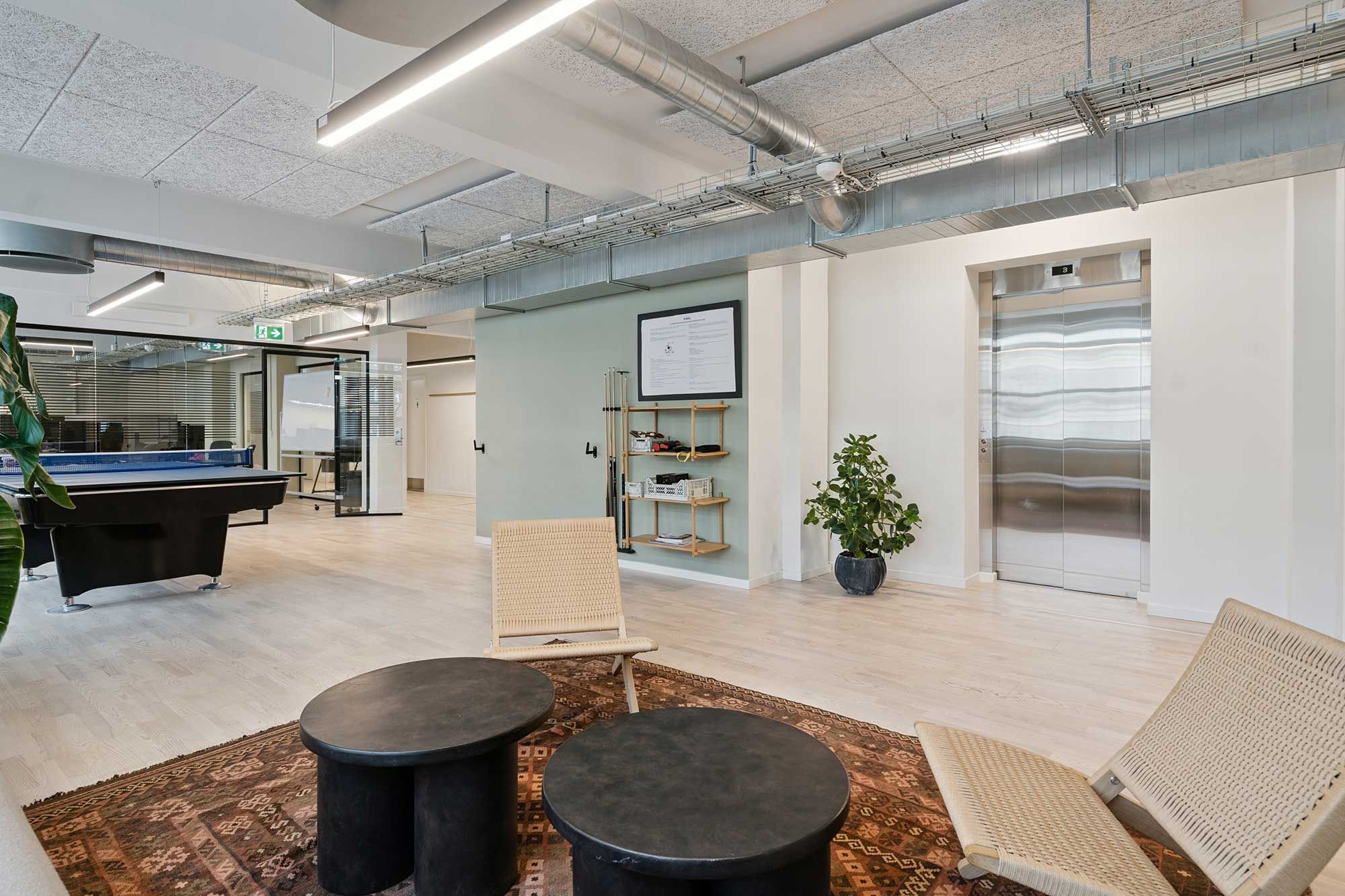 Kontorlokaler med lyse vægge og trægulve, træstole og bord, elevatordør, pingpongbord og plante i potte samt et skrift på væggen