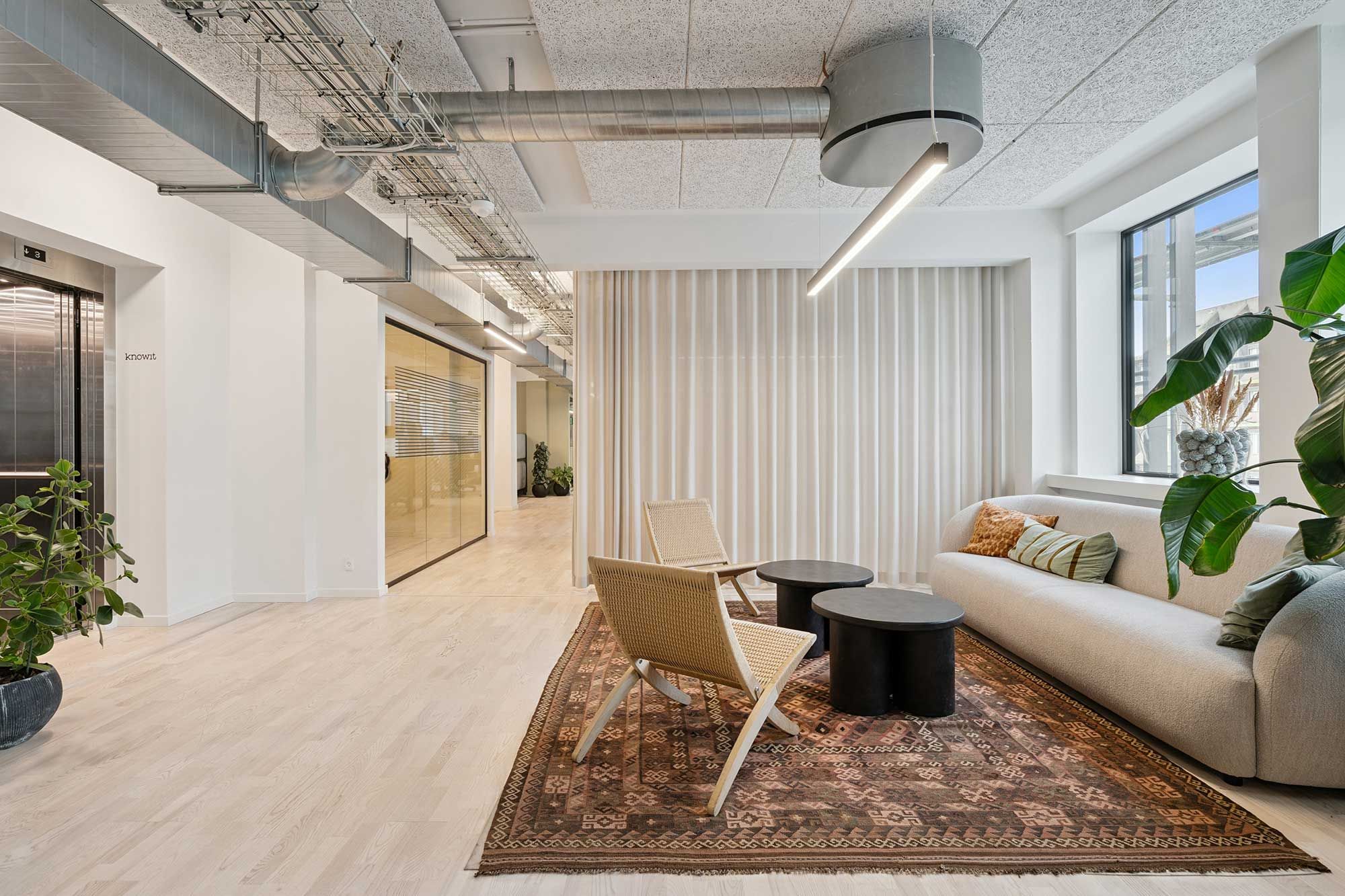 Inde i et moderne kontor med lyse lofter, hvide vægge og et stort vinduesparti, er der et hyggeligt opholdsområde med en sofa, en stol og to små kaffebord.