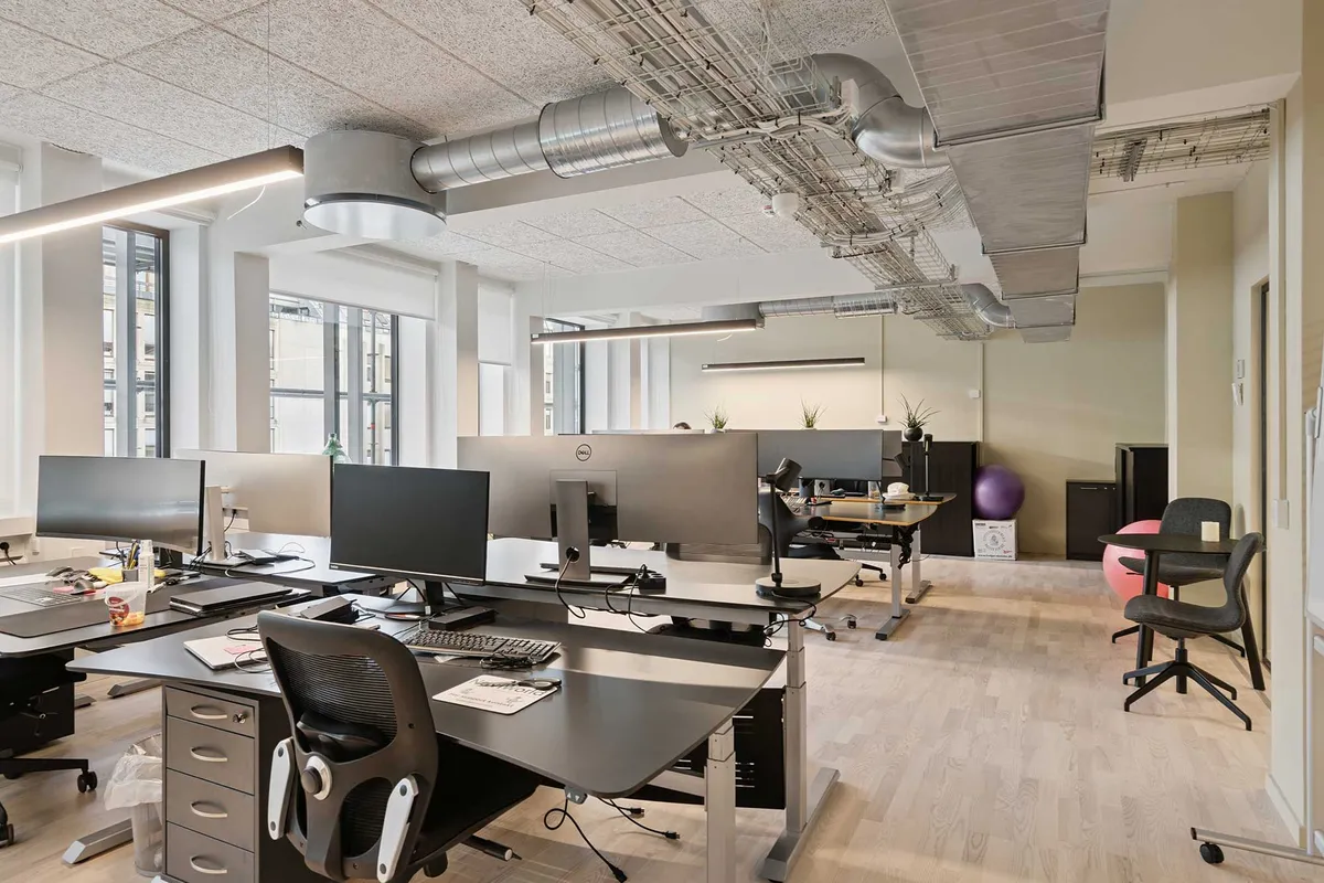 Kontor med moderne skriveborde, flere skærme og udstyr, høje lofter med ventilation, lyse loftslamper, og åbne vinduer med udsigt til byen