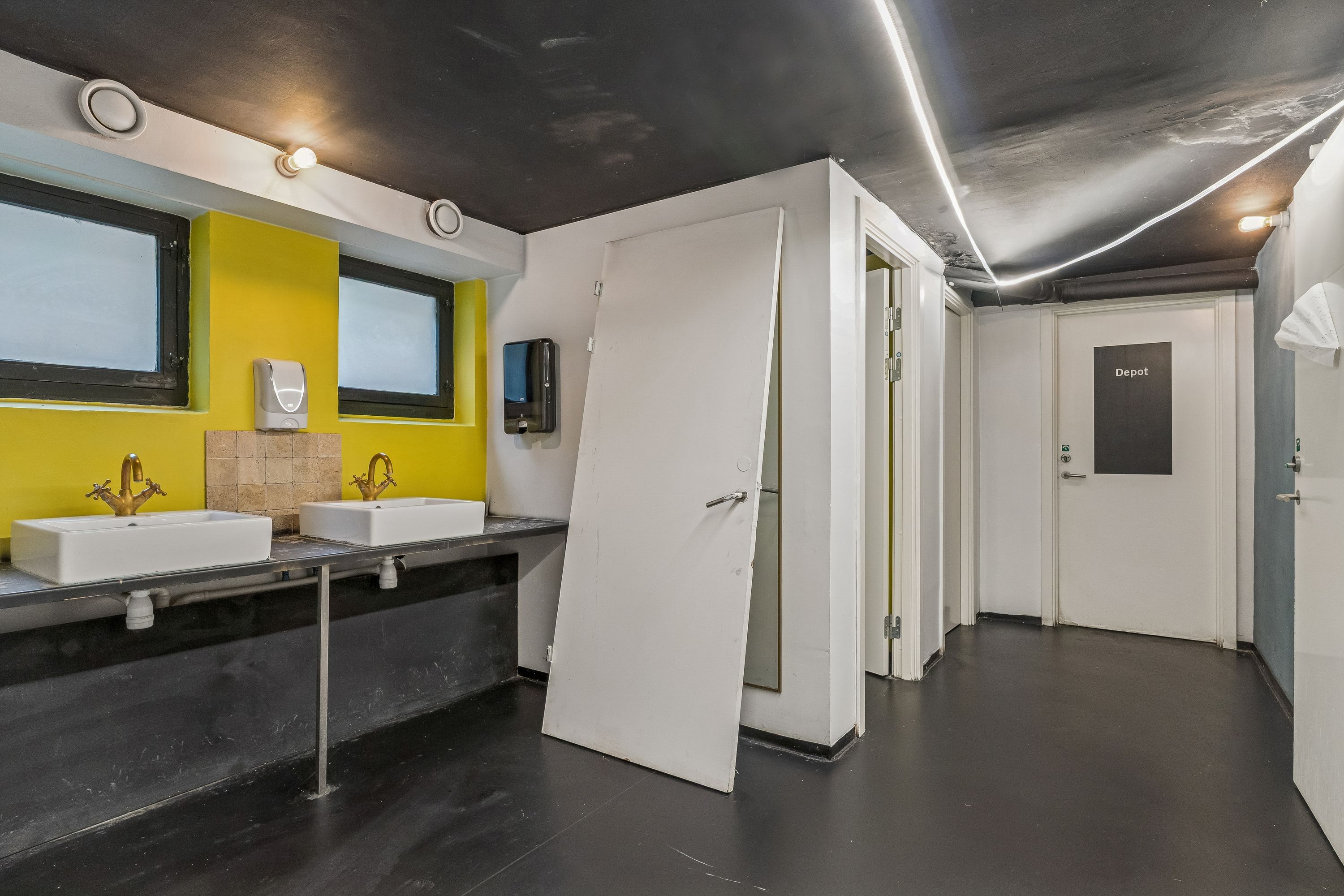 To toiletrum med hvide vasker og håndvask. Gule vægge med to vinduerne og to sæbeautomater. Lys pærer i loftet og to døre til toiletrum og depot.