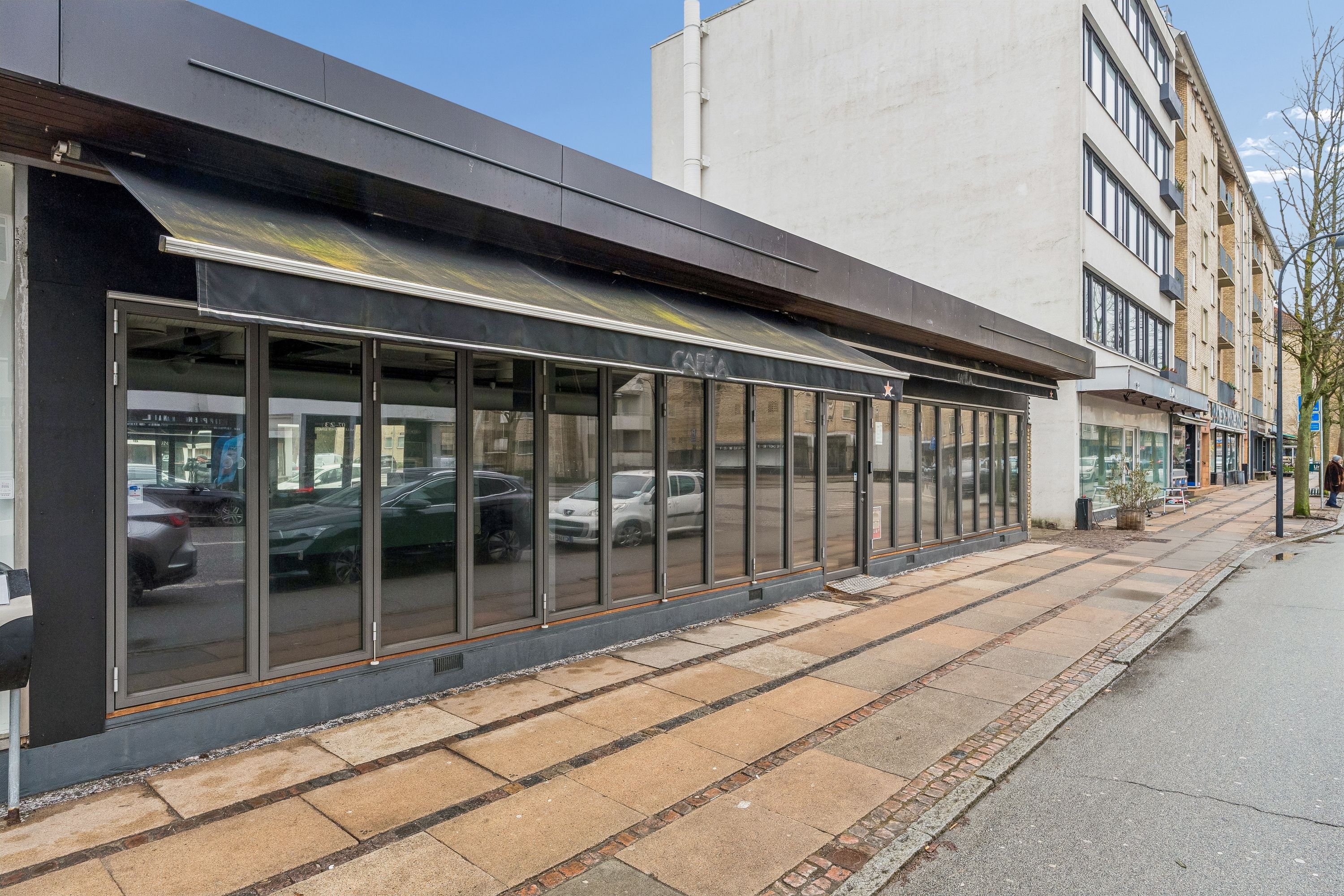 Moderne restaurant med store glasruder og sort aurbord på Ordrupvej 57 i Charlottenlund. Bagved står en bilparkering med flere biler og en person vandrer langs fortovet.