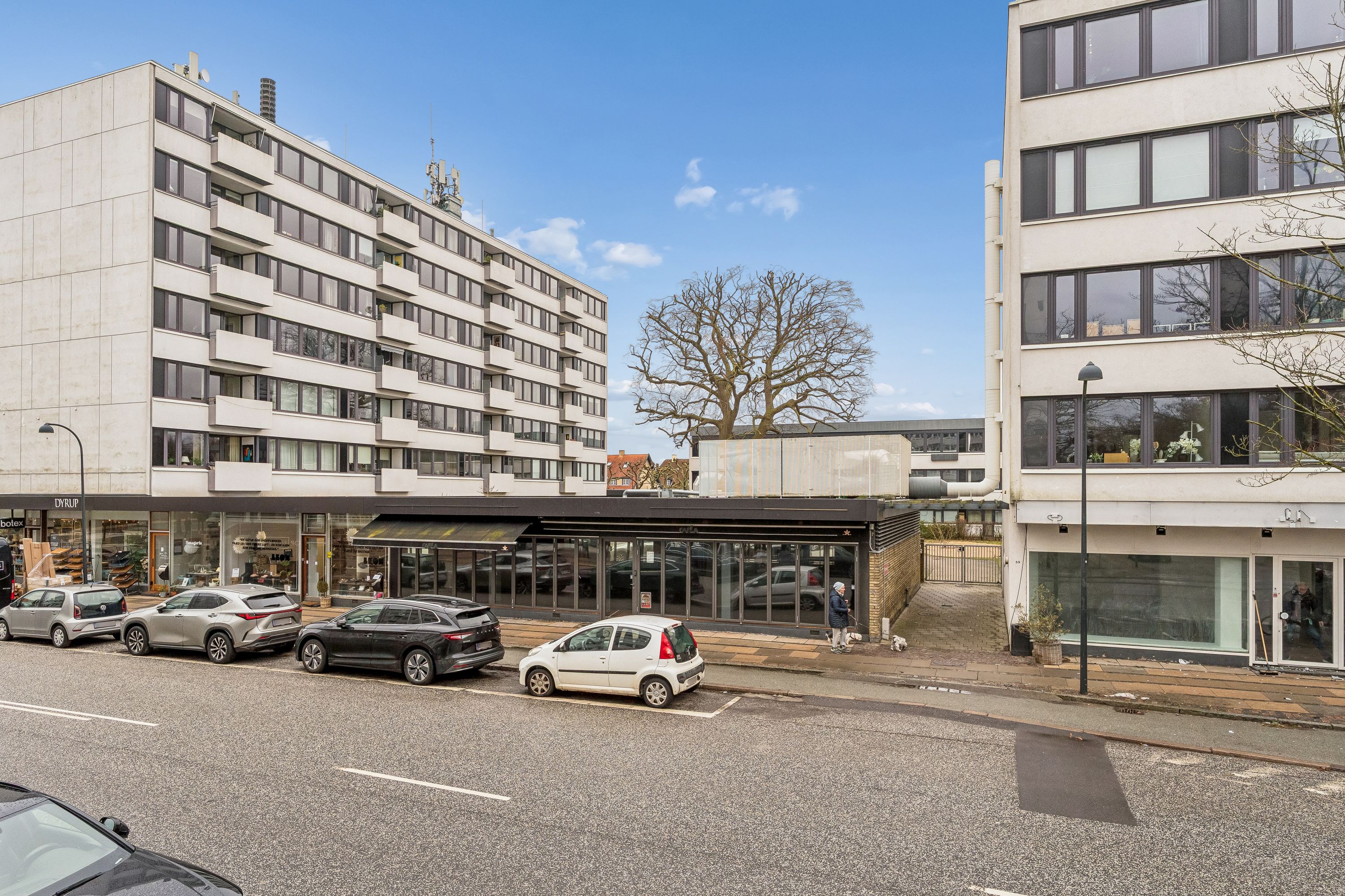 Søjle af beton med glasvinduer og balkoner og en restaurant med glasfront og skur i forgrunden og flere biler parkeret på gade og et gågadeområde med mennesker og planter