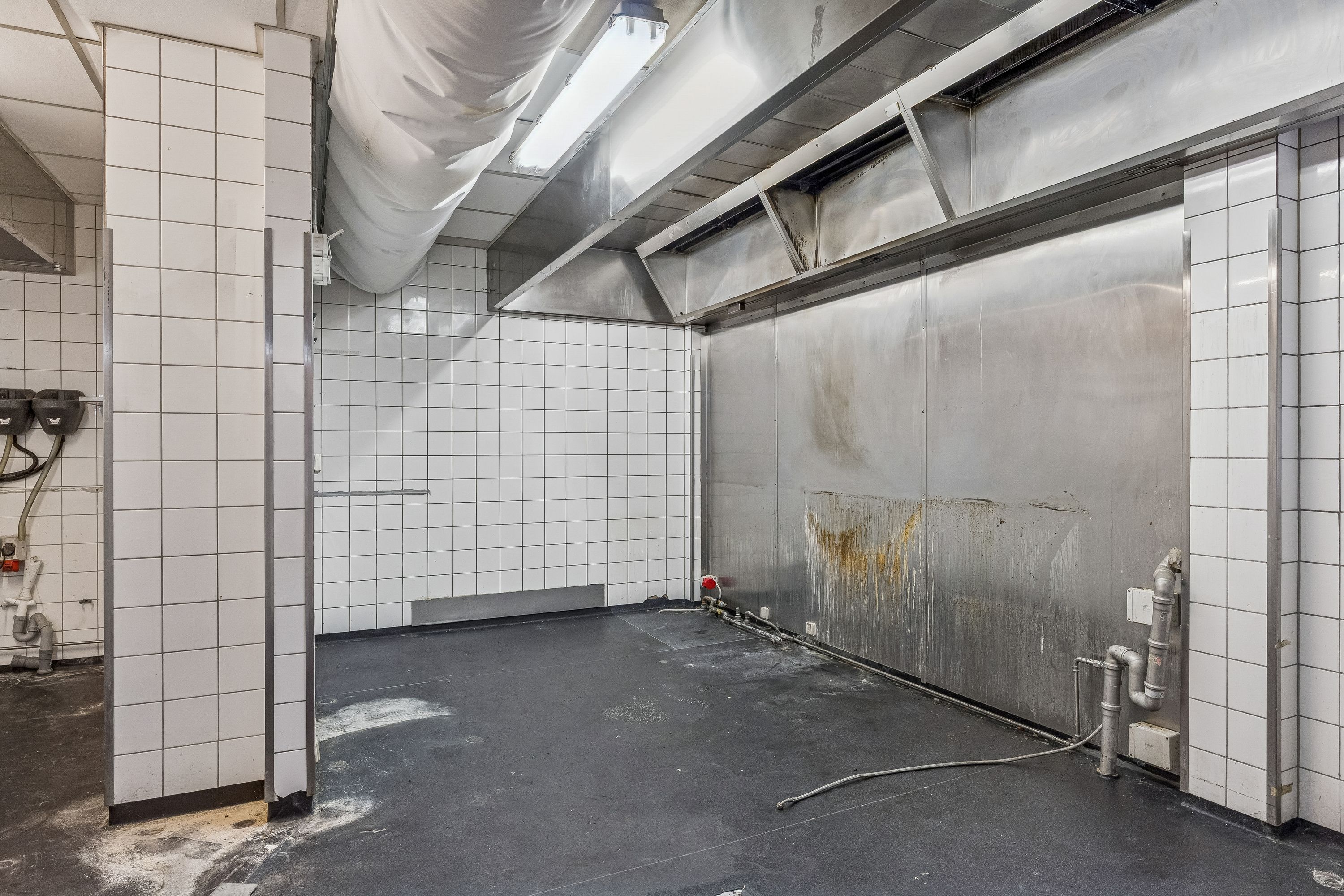 Et tomt køkken med hvide kakel og rustfrit stål. Stålbænk og ovn er forsynet med rust og rør. Et lysestage hænger fra loftet.