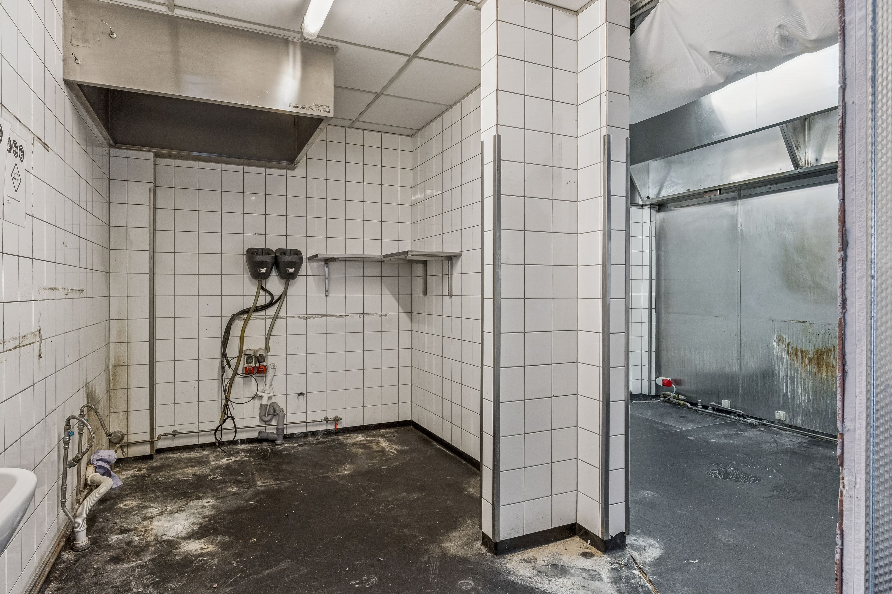 Restaurant til leje på Ordrupvej 57, Charlottenlund. En køkkenrum med hvide fliser, et spildevandsrør og to røde apparater.