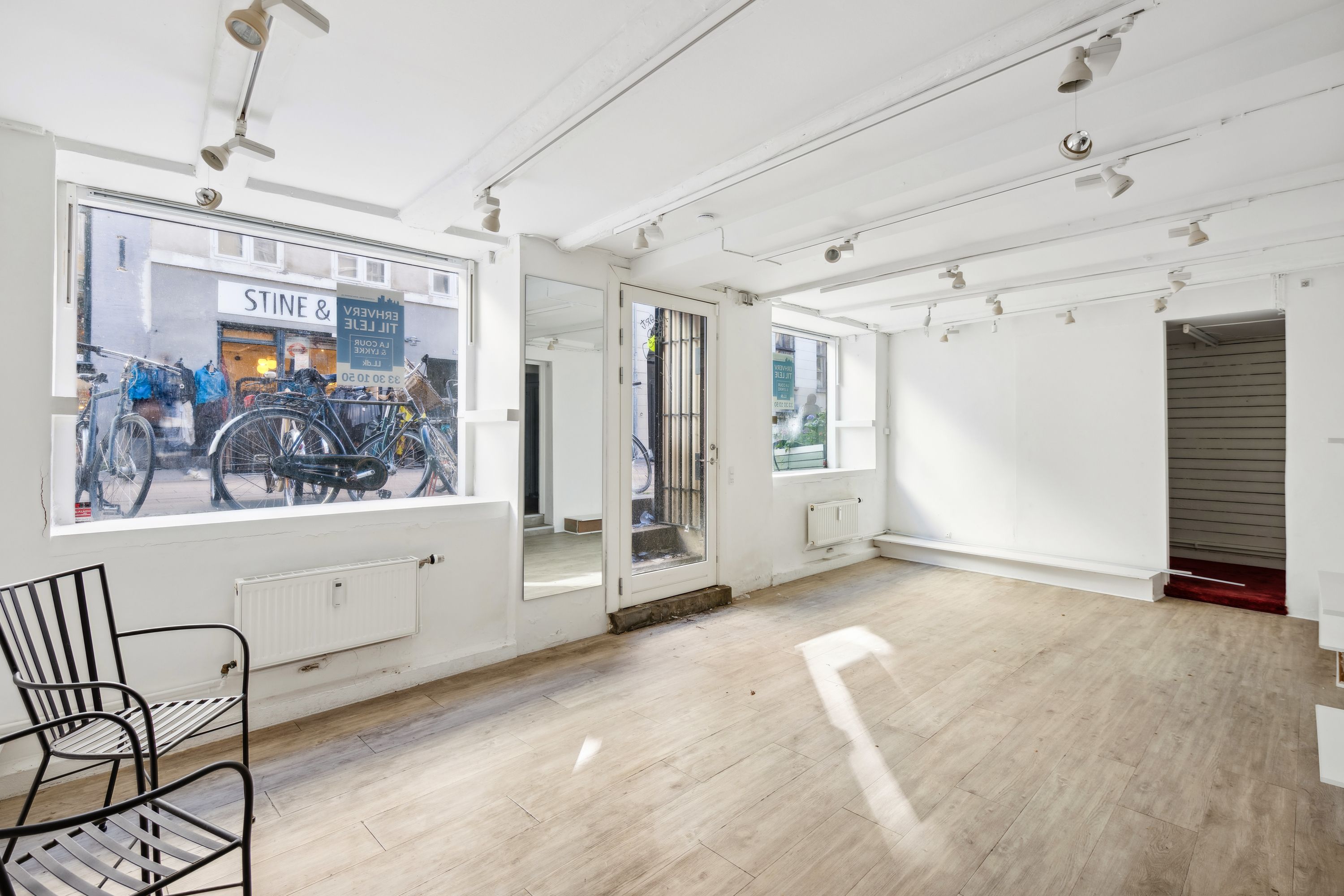 Butik med store ruder og lysarmatur. Et moderne møbel i sort og hvidt. En spejl og radiatorer på væggen. En stol og udgang til en anden butik