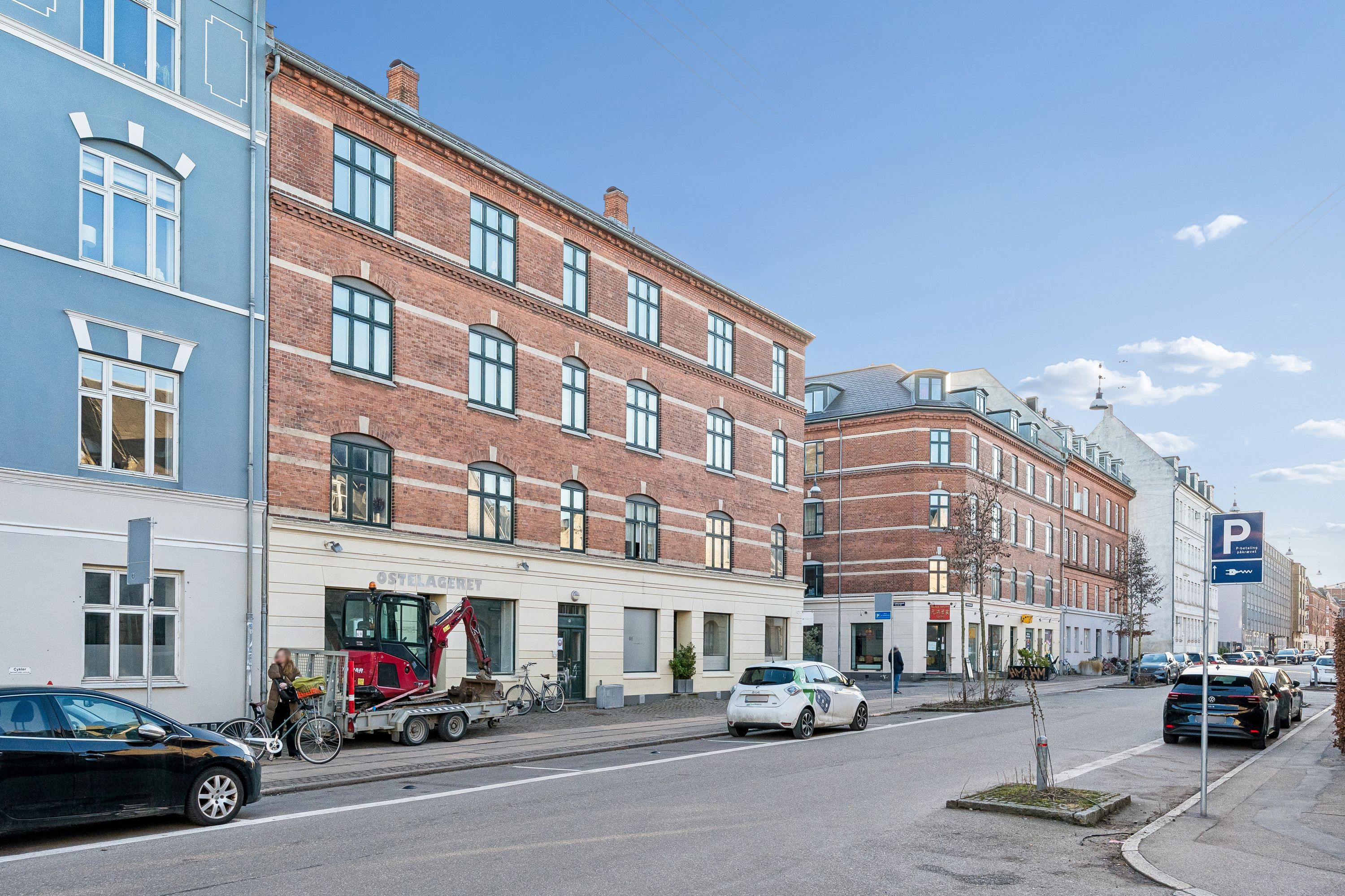 Mennesker parkerer biler på Landskronagade. En kvindelig person køber varer i en butik. En lastbil parkeret med en ekskavatermaskine på bagagerummet. En række husene og biler parkeret på siden af gaden.