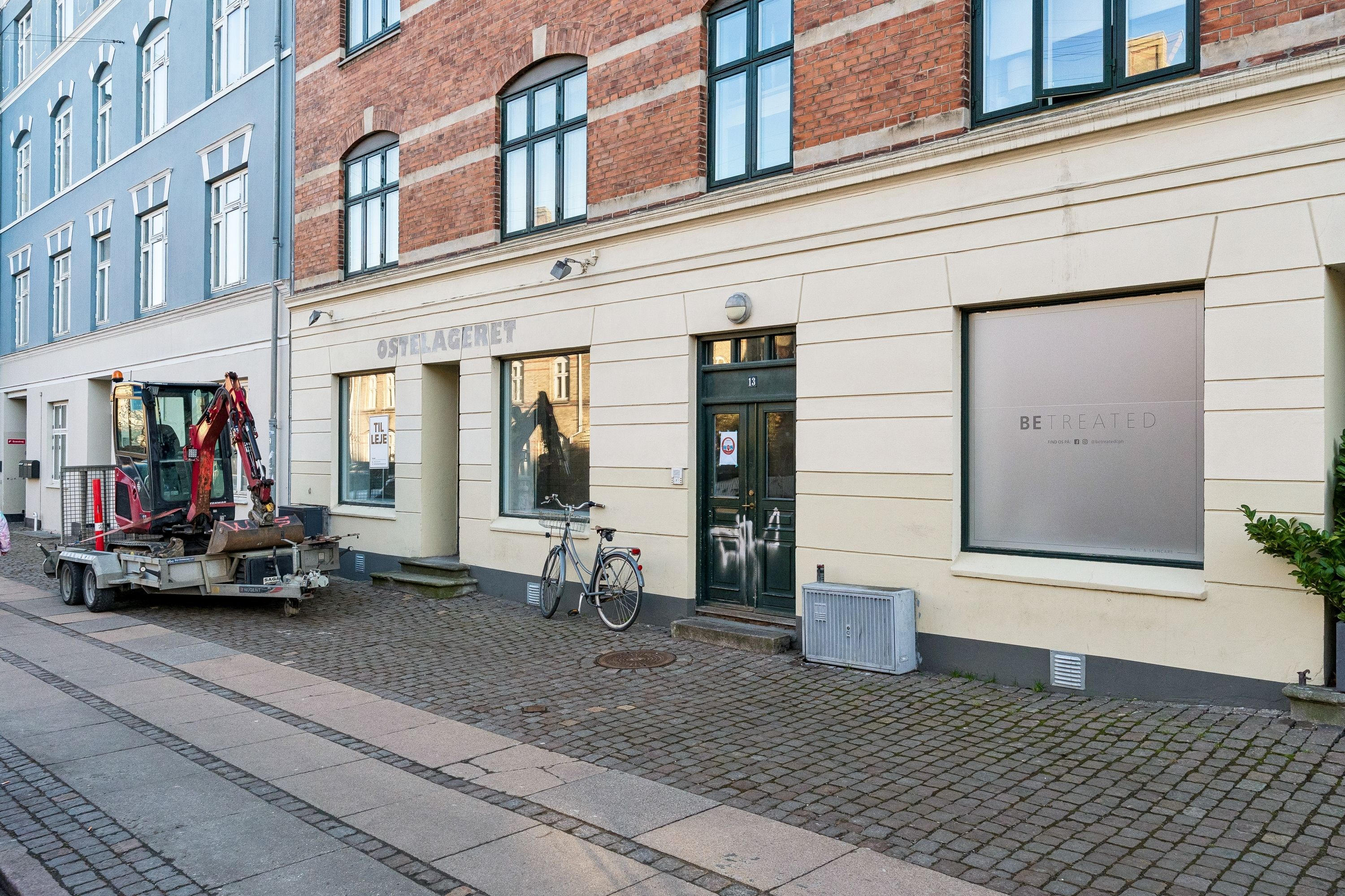 Butik til leje på Landskronagade 13 i København med ombygget facade og skilte med teksten Be Treated. En trækvogn med gravemaskine og cykel står foran bygningen