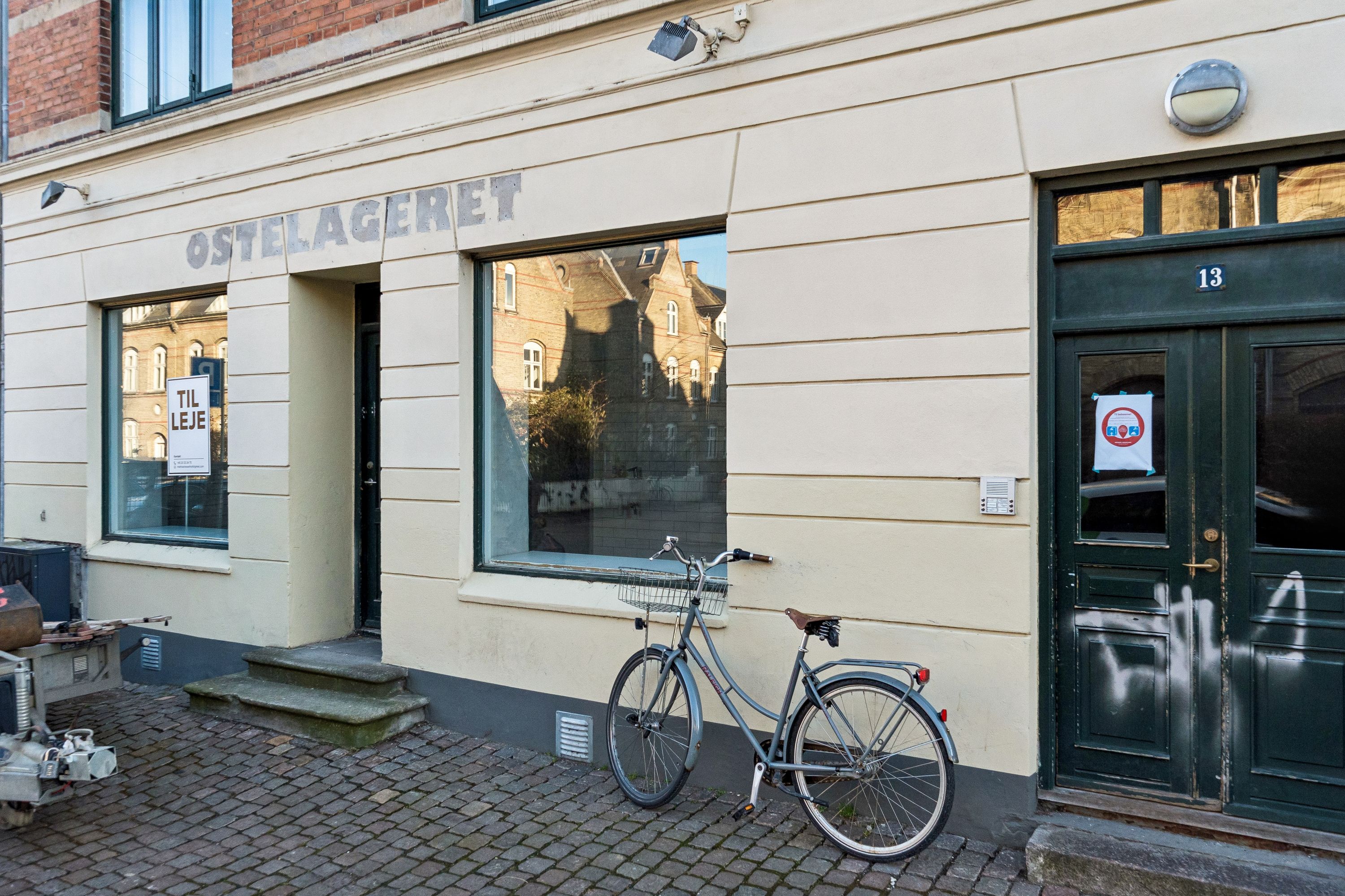 En cykel står foran en butik med teksten Ostelageret på facaden. Der er en skrift, der siger Til Leje, samt en dør med nummer 13. Der er graffiti på døren og spor af graffiti på væggen.