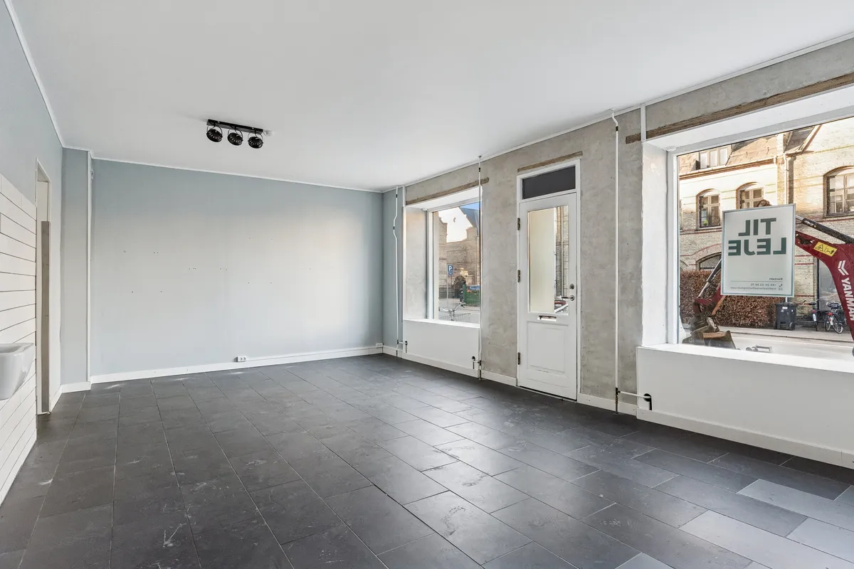 Moderne, tom lejemål med glasvinduer, hvide vægge, sort gulv og tre spots i loftet. Butikken er til leje og har reklameskilt med teksten Leje.