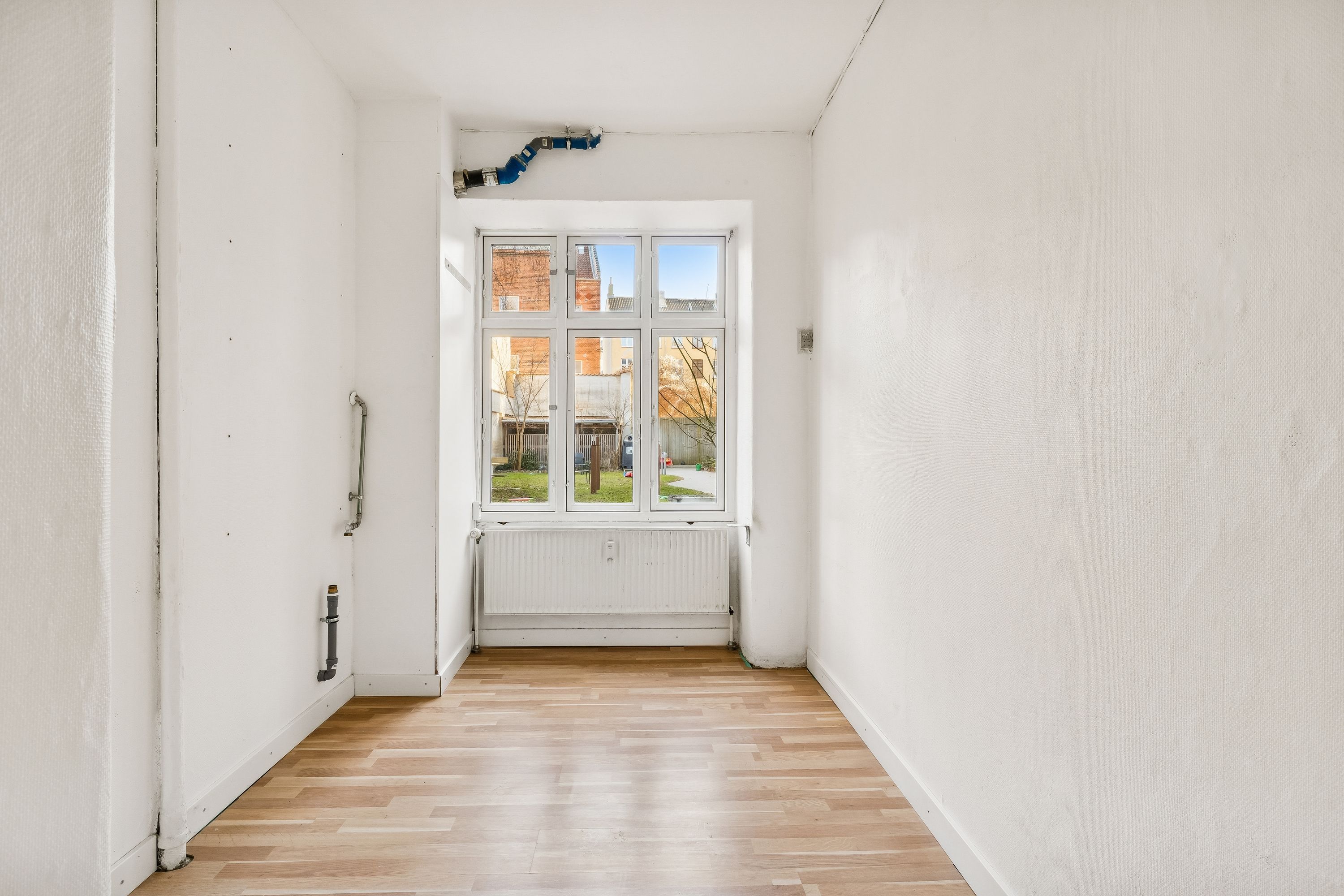 Langt og mørkt korridor med hvid væg og gulvtæppe samt vindue med udsigt til grønne områder og bygninger på Landskronagade i København